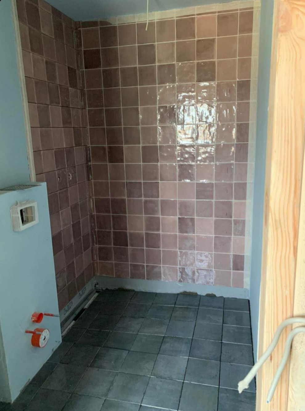 Wykończona łazienka z brązowymi płytkami na ścianach i szarymi na podłodze. Widoczne elementy instalacji wodnej i fragment drewnianej konstrukcji.