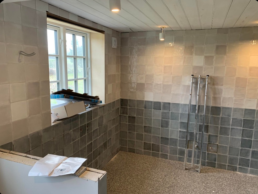 Remont łazienki w trakcie: ułożone kwadratowe płytki w dwóch odcieniach szarości, drabina oparta o ścianę, okno z materiałami budowlanymi, dokumentacja na murku.