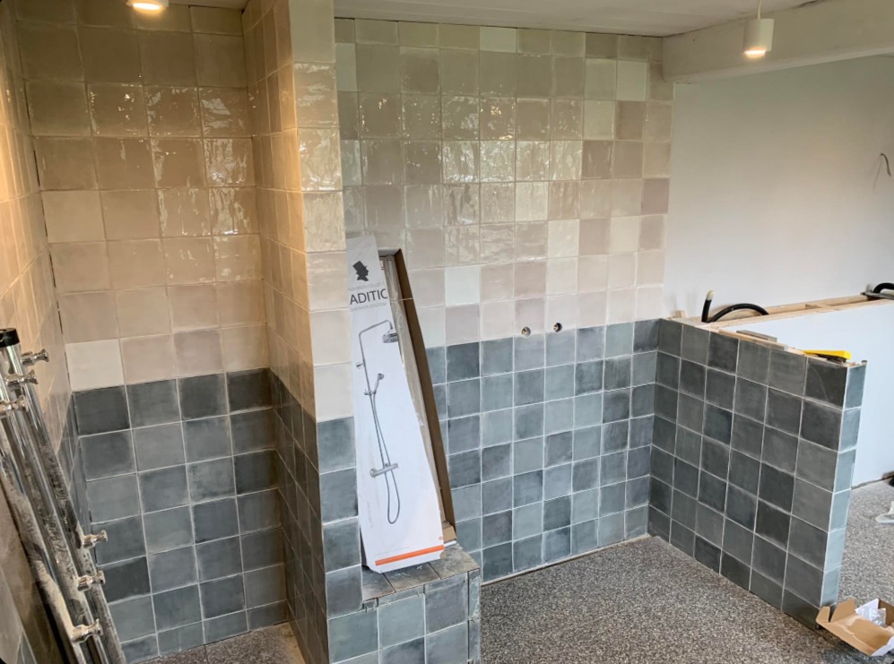 Łazienka w trakcie wykańczania: ułożone kwadratowe płytki w odcieniach szarości i beżu, widoczna drabinka i karton z zestawem prysznicowym.