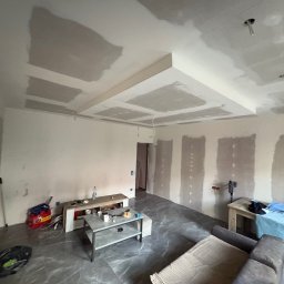 REMONTERS DAMIAN MAŁECKI - Pokój w trakcie remontu: szpachlowane ściany i sufit z dekoracyjną zabudową gipsową. Widoczne narzędzia i materiały budowlane. Podłoga wyłożona szarymi płytkami imitującymi marmur.