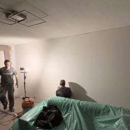 Dwóch mężczyzn podczas prac wykończeniowych w pomieszczeniu. Jeden wchodzi, drugi szpachluje ścianę. Widoczne narzędzia, lampa i zabezpieczone meble folią.