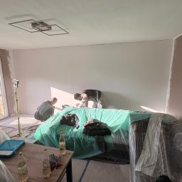 REMONTERS DAMIAN MAŁECKI - Dwóch mężczyzn szpachluje ścianę w jasnym pokoju, zabezpieczony mebel pod folią, widoczne narzędzia i oświetlenie. Ściana częściowo w kolorze.