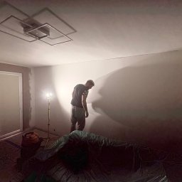 REMONTERS DAMIAN MAŁECKI - Pokój w trakcie prac wykończeniowych: ściana po nałożeniu gładzi, lampa budowlana, zakryte folią meble. Widoczny pracownik w trakcie pracy.