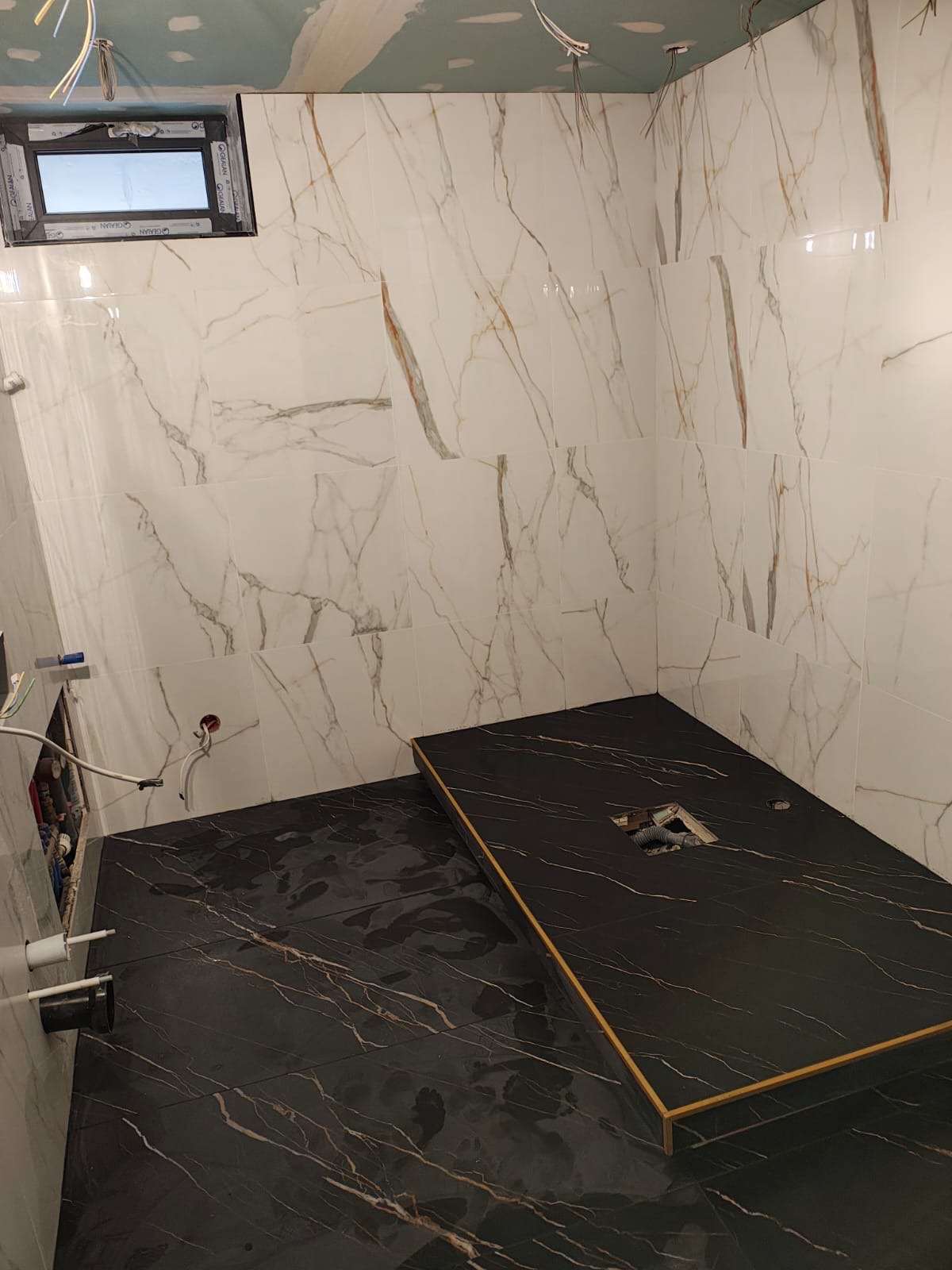 Wnętrze łazienki w trakcie prac wykończeniowych: białe płytki marmurowe na ścianach, czarna podłoga z płytek imitujących marmur, widoczne instalacje i sufit z płyt karton-gips.