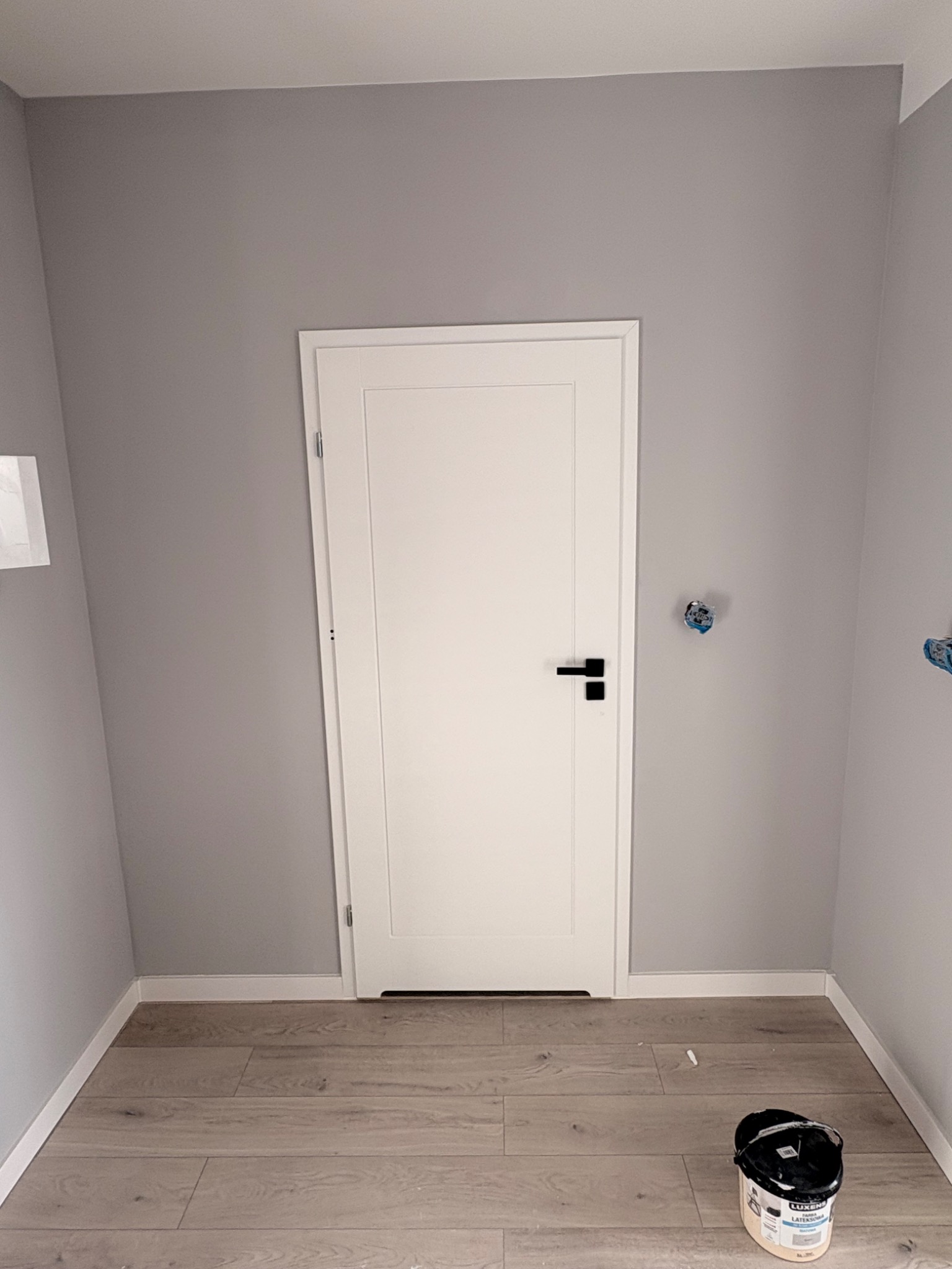 Szara ściana z białą, zamkniętą stolarką drzwiową i drewnopodobną podłogą. Widoczne puszki elektryczne i wiadro z farbą. Minimalistyczne wykończenie wnętrza.