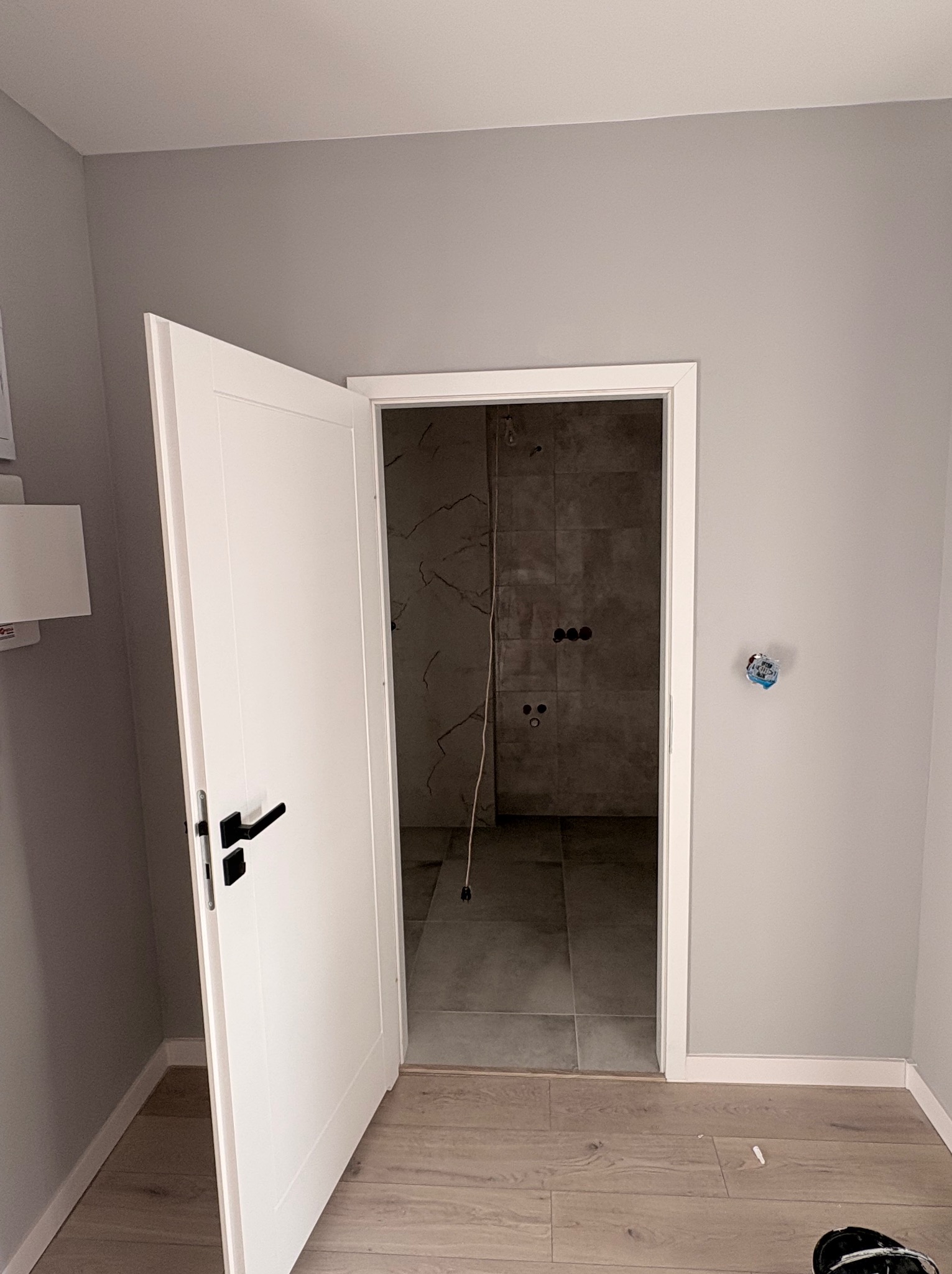 Otwarta biała, minimalistyczna  drzwi z czarną klamką, prowadząca do wykończonej łazienki z szarymi płytkami. Widoczne przygotowane punkty elektryczne.