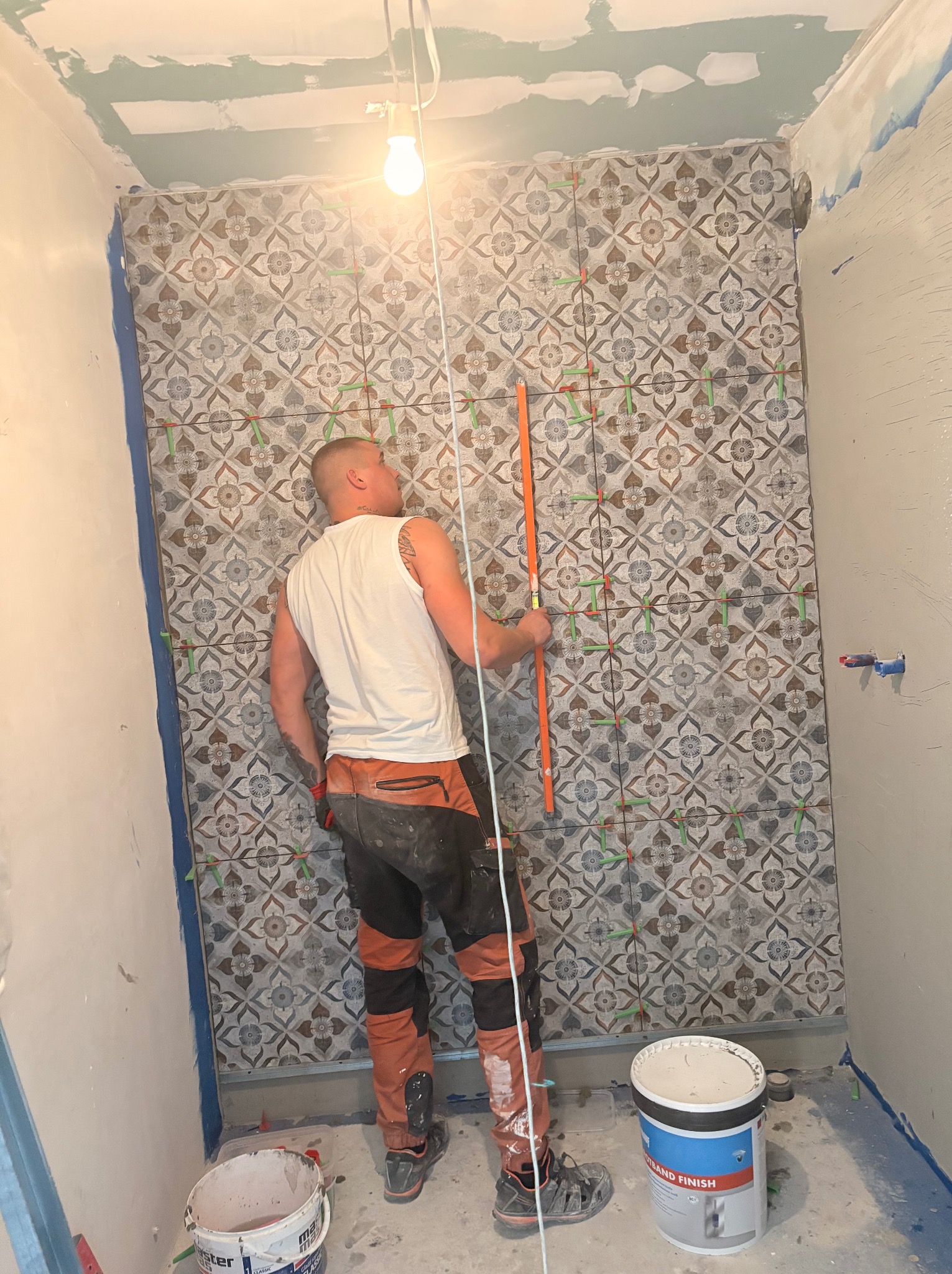 Układanie płytek wzorzystych na ścianie w łazience. Glazurnik z poziomicą, widoczne krzyżyki dystansowe i wiadro z zaprawą. Ściany przygotowane do dalszych prac.