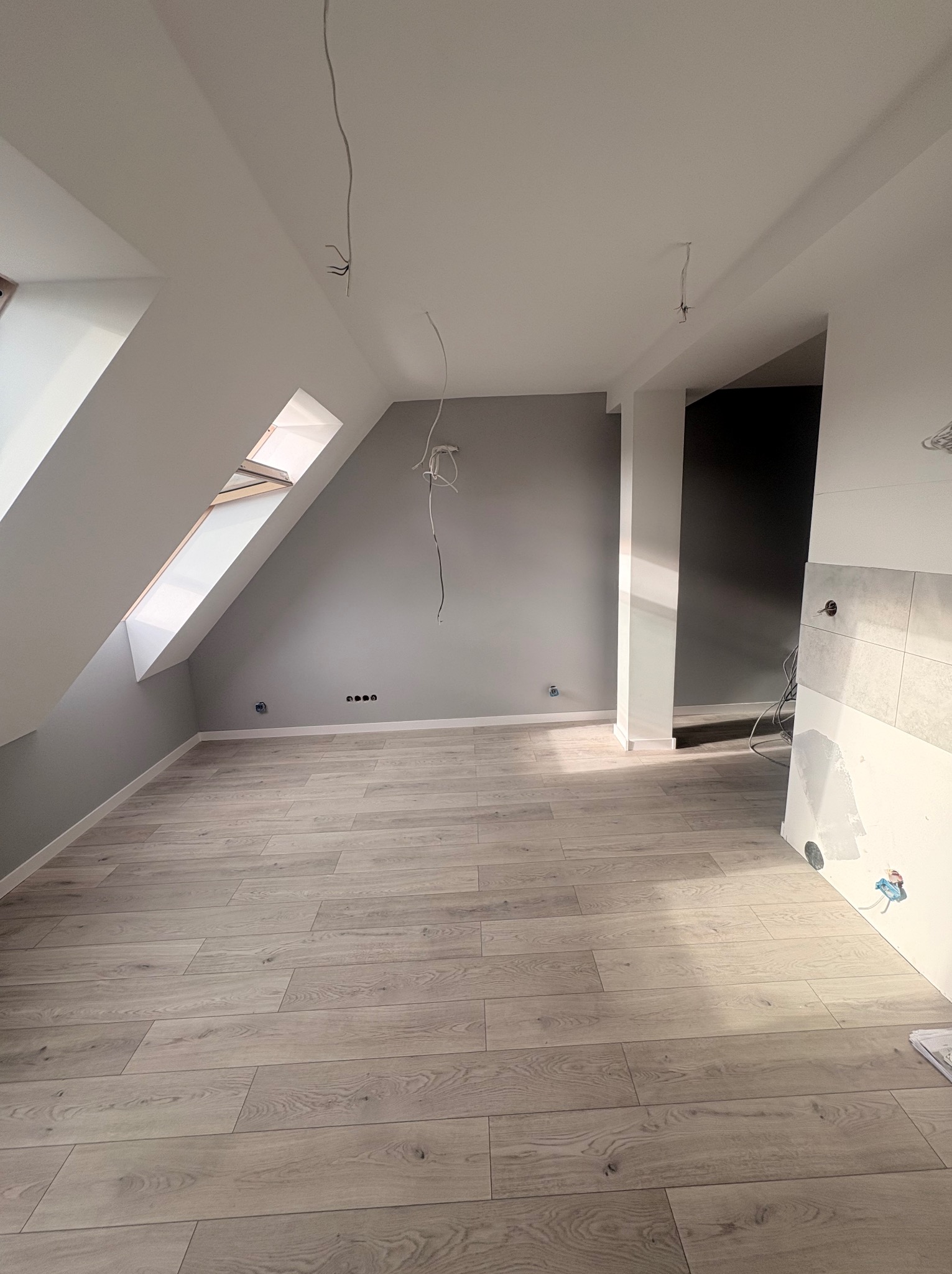 Jasne panele podłogowe w pokoju na poddaszu z oknem dachowym. Szara ściana z gniazdkami, częściowo wykończona ściana z płytkami. Wiszące kable elektryczne.