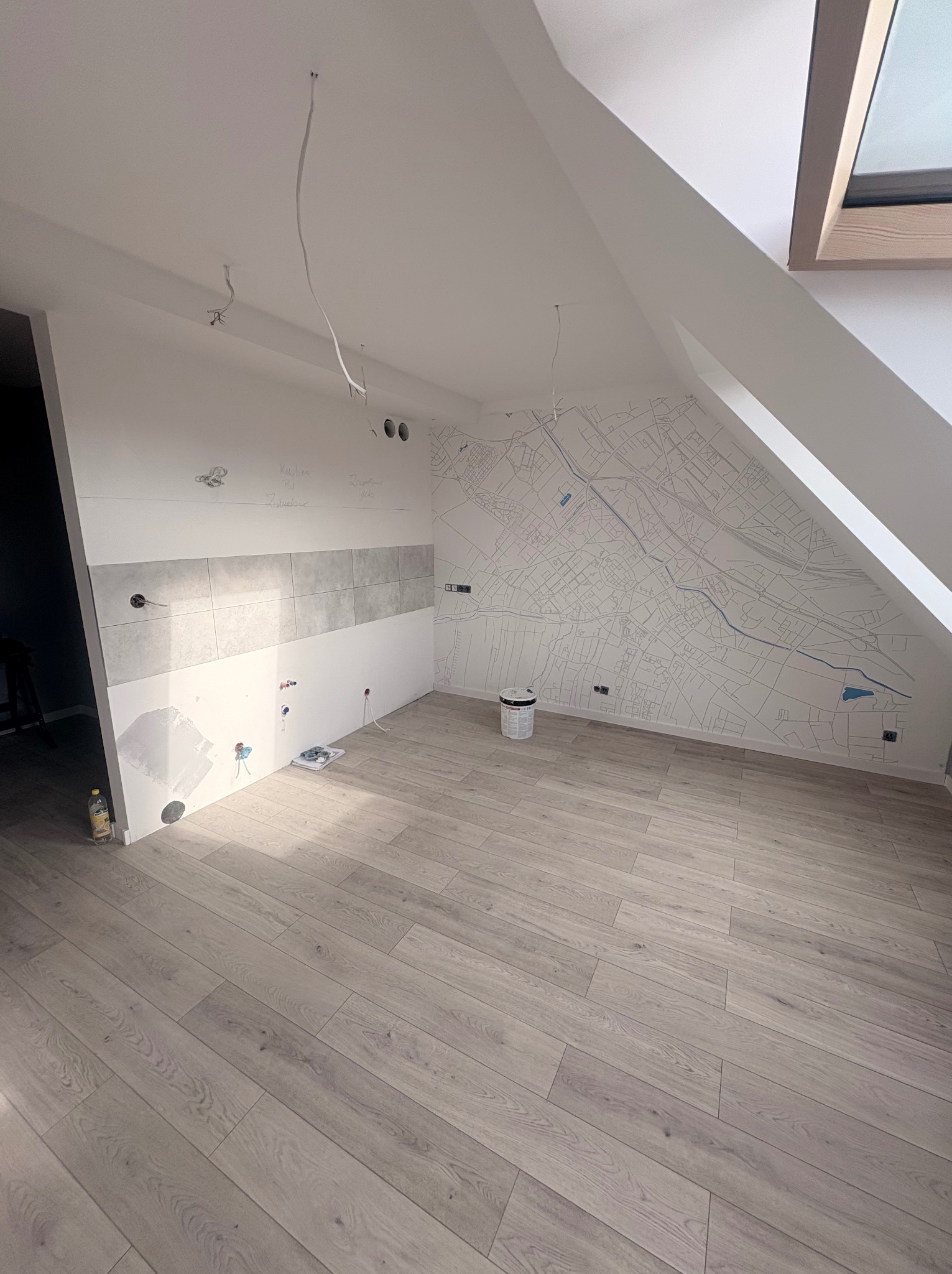 Wnętrze poddasza w trakcie wykończenia: ułożona podłoga z paneli, częściowo położone płytki na ścianie, na jednej ze ścian mapa miasta. Widoczne kable i puszki elektryczne.