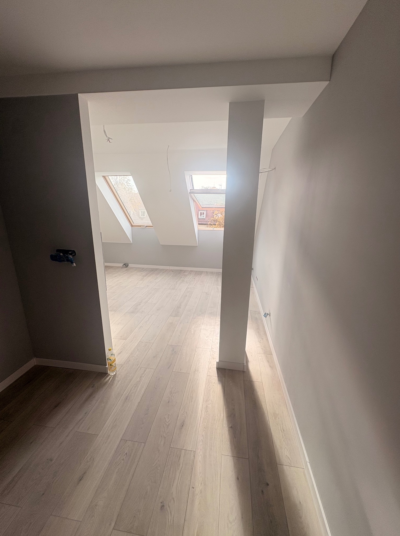 Jasne poddasze po remoncie z drewnianą podłogą i oknami dachowymi. Świeżo pomalowane ściany w odcieniach szarości. Widoczne gniazdka elektryczne i wystające przewody.