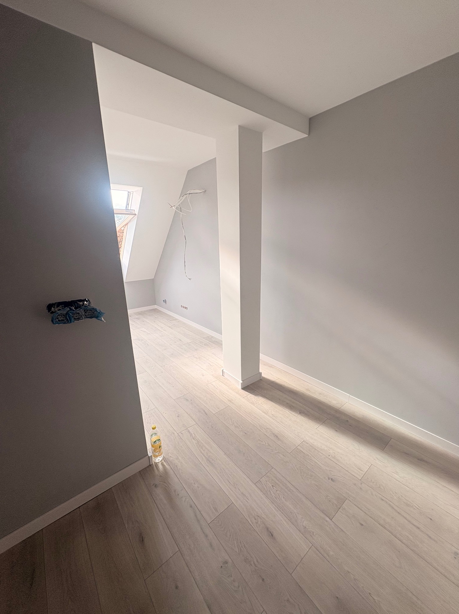 Jasne panele podłogowe w szarym pokoju na poddaszu z oknem dachowym. Widoczne kable elektryczne i puszka instalacyjna. Butelka z napojem przy ścianie.