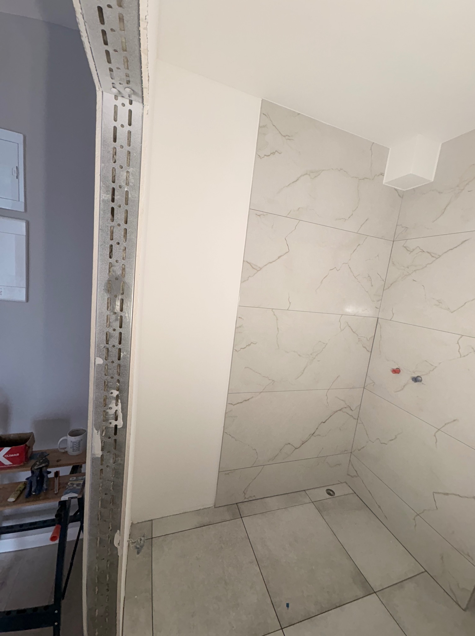 Fragment łazienki w trakcie prac: ułożone płytki imitujące marmur na ścianie i podłodze, widoczny profil metalowy przy ścianie, przygotowania do dalszych prac.