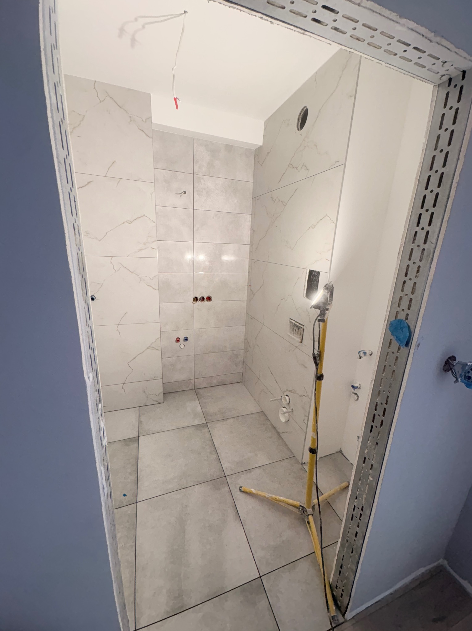 Wnętrze łazienki w trakcie wykańczania: ułożone płytki imitujące marmur na ścianach i podłodze, widoczne instalacje, oświetlenie robocze i metalowe profile przy drzwiach.