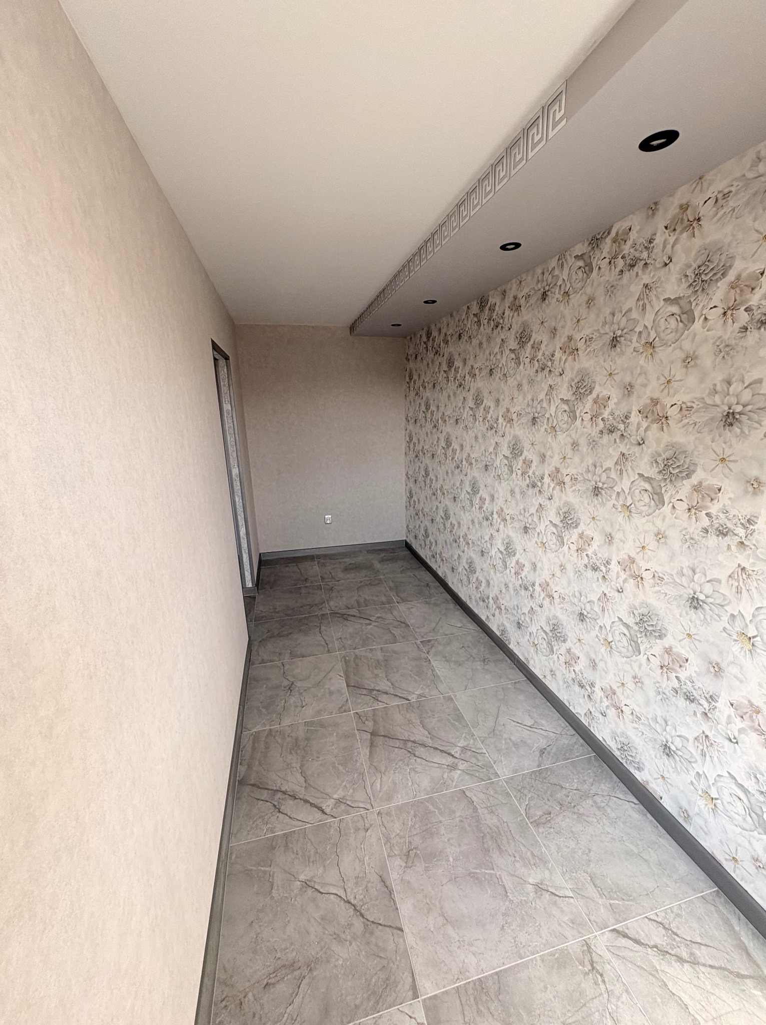 Wykończone wnętrze: szare płytki podłogowe imitujące marmur, ściana z tapetą w kwiaty, listwa dekoracyjna pod sufitem z oświetleniem punktowym.