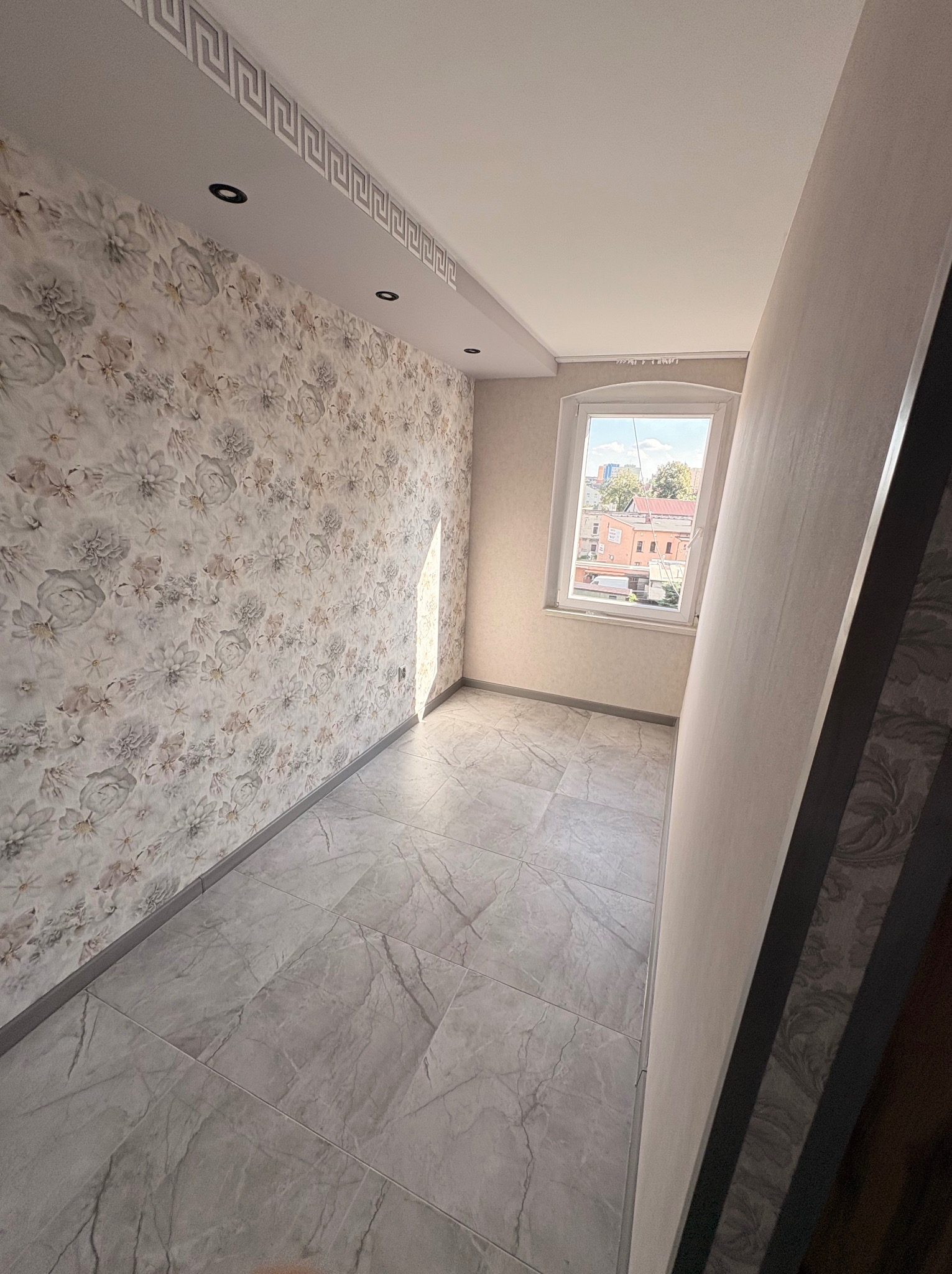Wykończone wnętrze pokoju z tapetą w kwiaty, szarą podłogą z płytek i oknem z widokiem na budynki. Dekoracyjny pasek na suficie. Nowoczesne wykończenie.