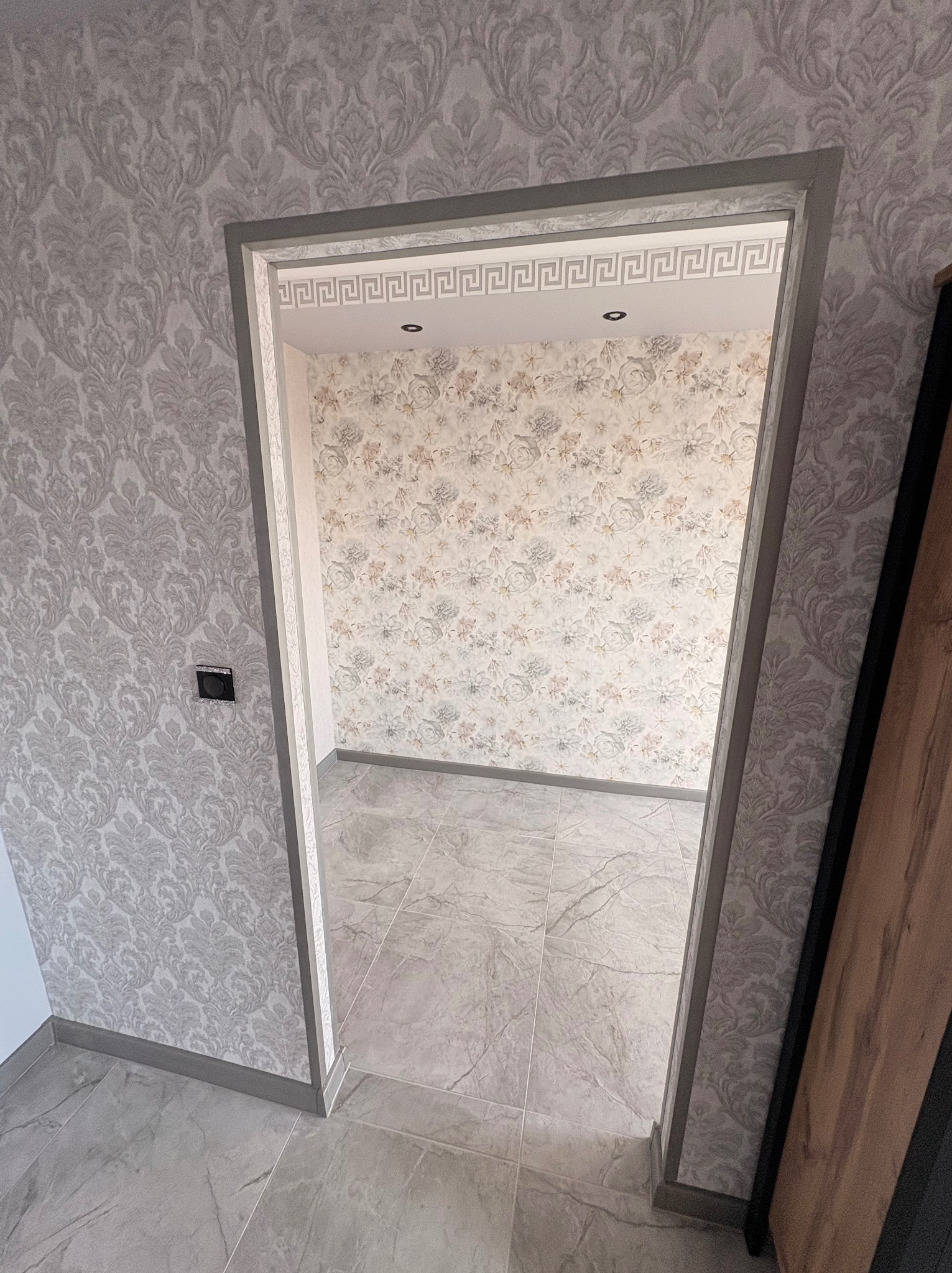 Wykończone wnętrze z wzorzystą tapetą i szarą ramą drzwi z motywem greckim. Podłoga wyłożona płytkami imitującymi marmur. Widok z perspektywy.