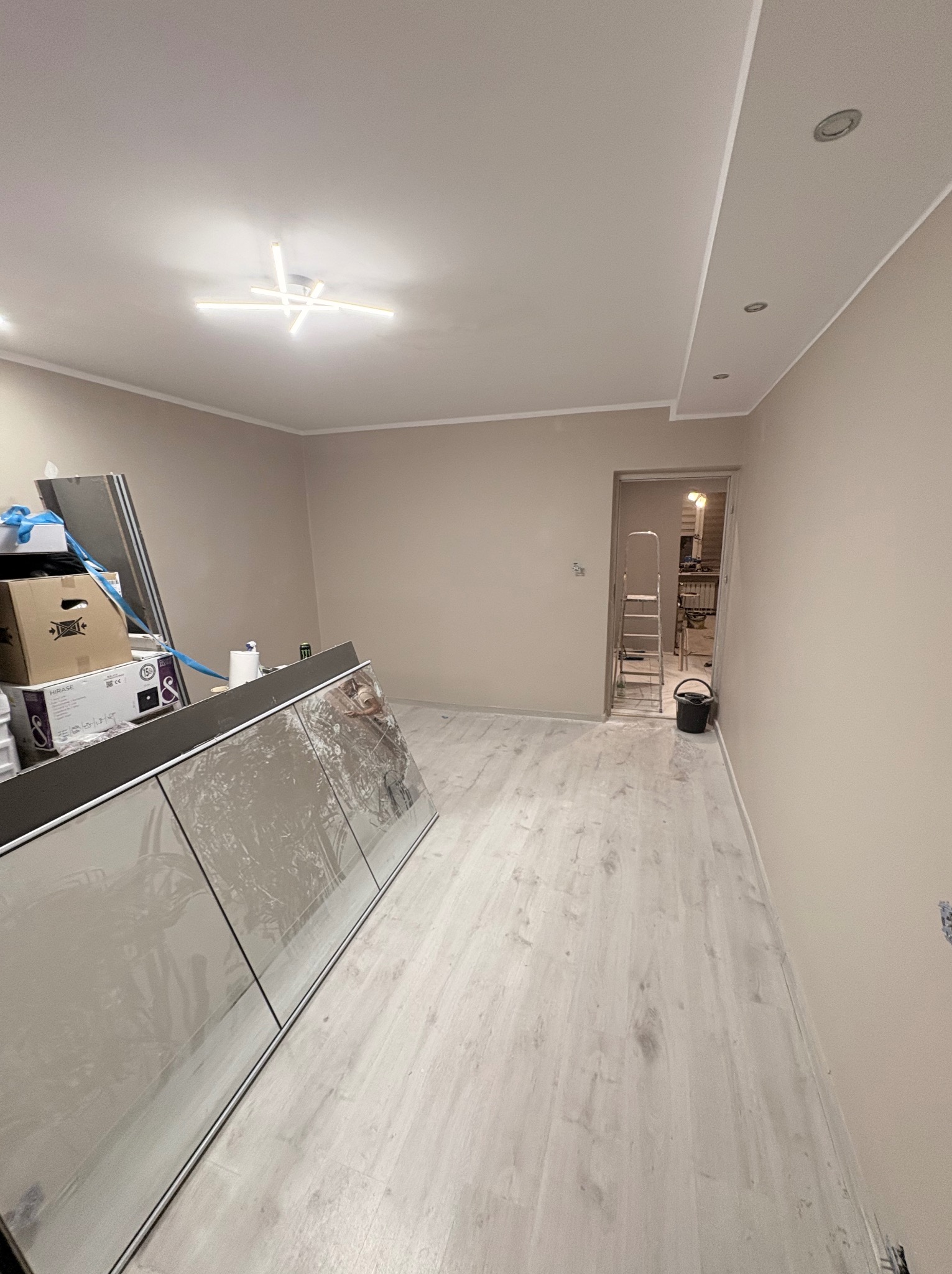 Jasne wnętrze w trakcie wykańczania: pomalowane ściany w odcieniach beżu, panele podłogowe, widoczne elementy zabudowy kuchennej i drabina malarska w przejściu.