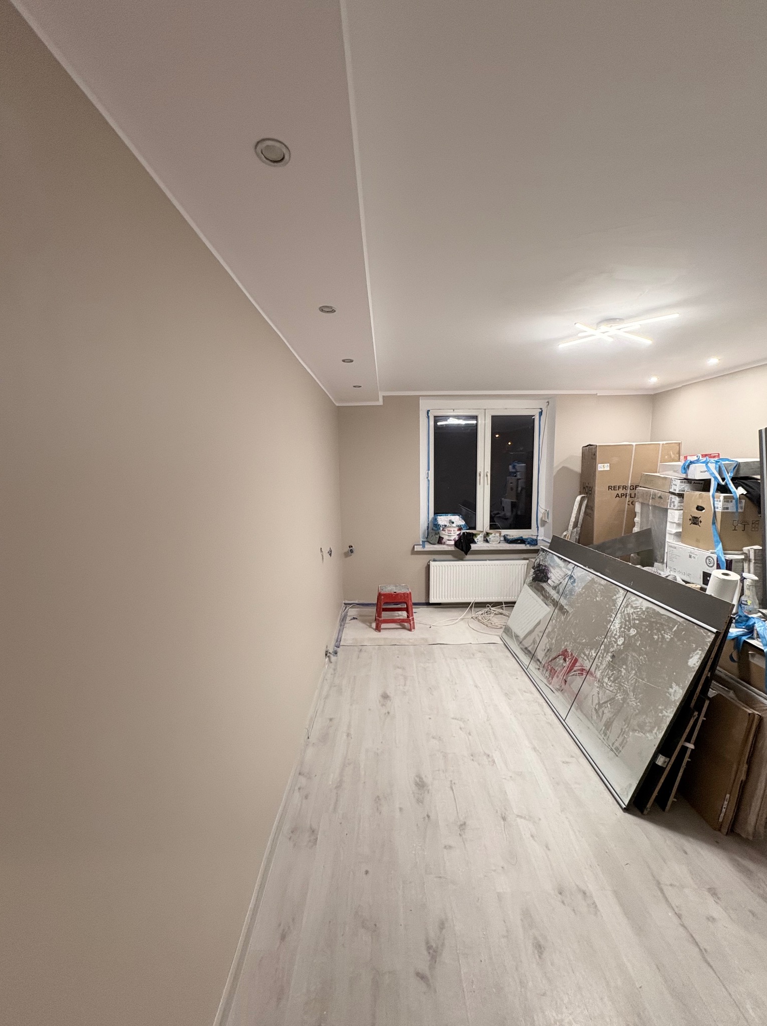 Jasne wnętrze w trakcie prac wykończeniowych: pomalowane ściany, nowe panele podłogowe, okno z grzejnikiem i sterta materiałów budowlanych. Widoczne punkty oświetleniowe.