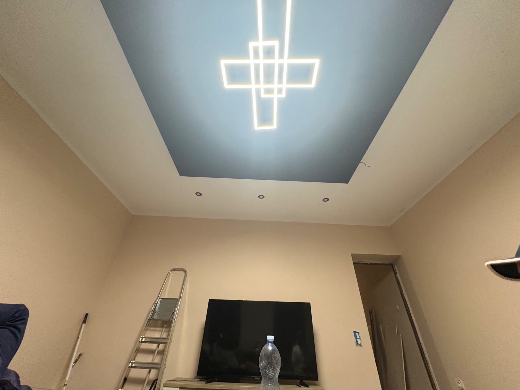 Nowoczesny sufit podwieszany z geometrycznym oświetleniem LED w kształcie krzyża, w pokoju z beżowymi ścianami i drabiną opartą o ścianę.