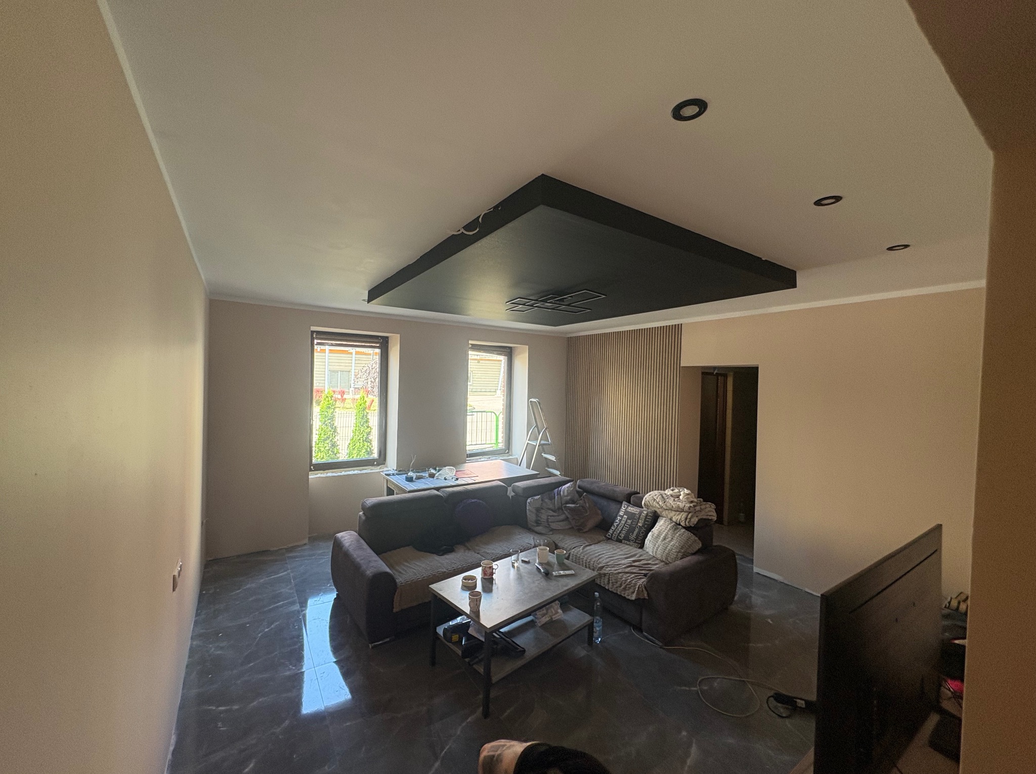 Salon po malowaniu ścian na beżowo z czarnym podwieszanym sufitem i marmurową podłogą. Widoczne okna i częściowo zasłonięty narożnik z stolikiem kawowym.