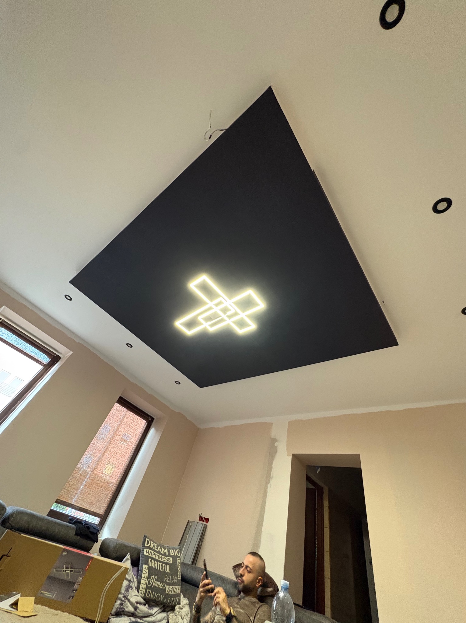 Nowoczesny sufit podwieszany z czarnym panelem i geometrycznym oświetleniem LED w kształcie krzyża. Widoczne okna dachowe i fragment wnętrza w trakcie wykończenia.