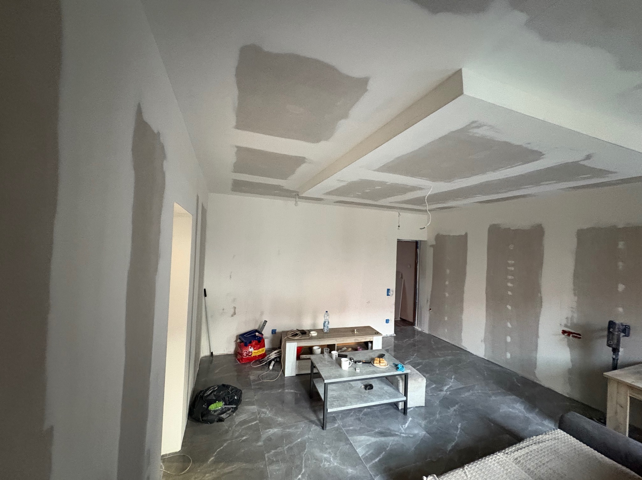 Pokój w trakcie szpachlowania: ściany i sufit z widocznymi łatami gipsowymi, szare płytki na podłodze, meble w tle. Widoczne narzędzia i materiały budowlane.
