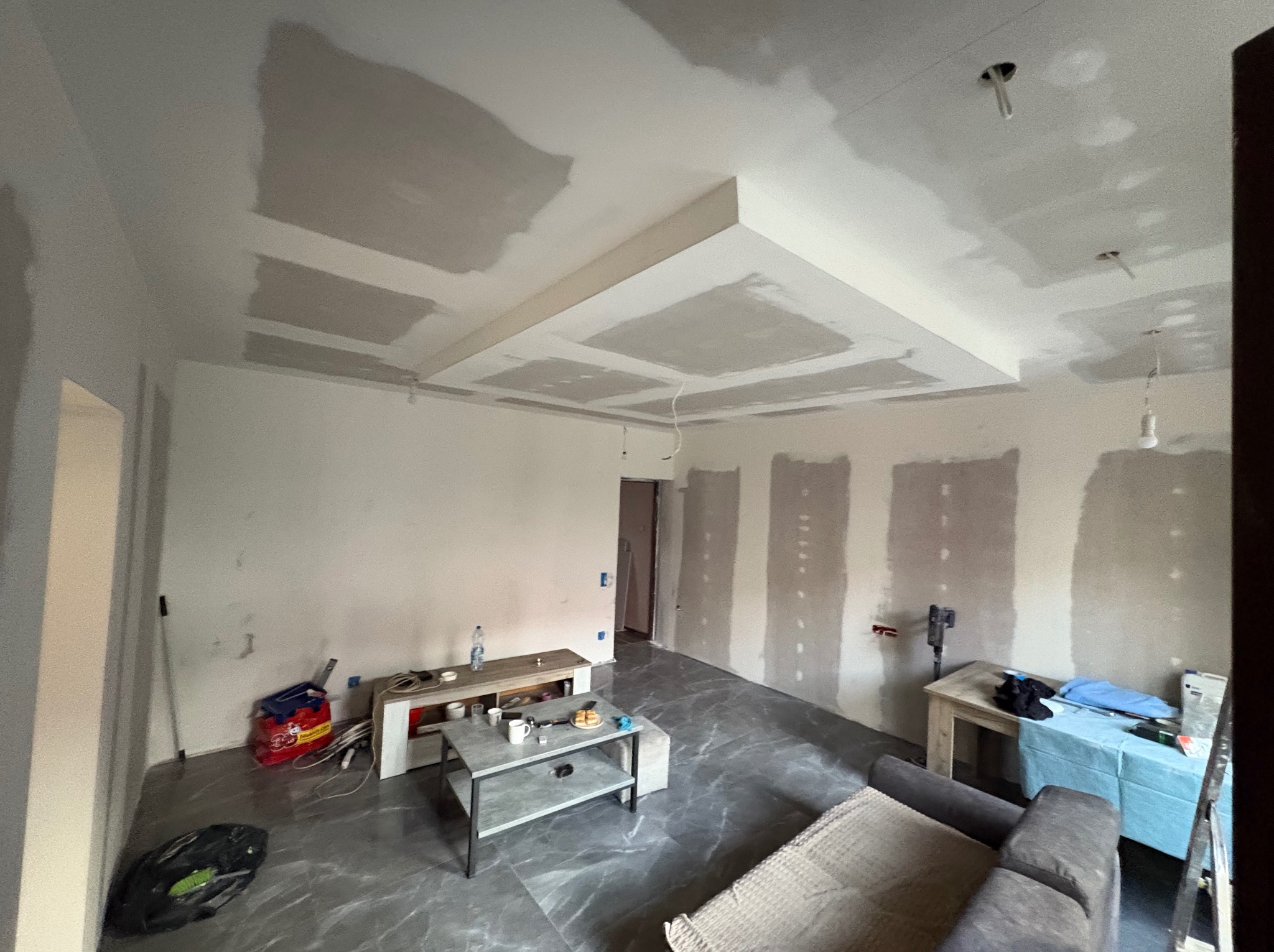 Pokój w trakcie remontu: szpachlowane ściany i sufit z dekoracyjną zabudową gipsową. Widoczne narzędzia i materiały budowlane. Podłoga wyłożona szarymi płytkami imitującymi marmur.