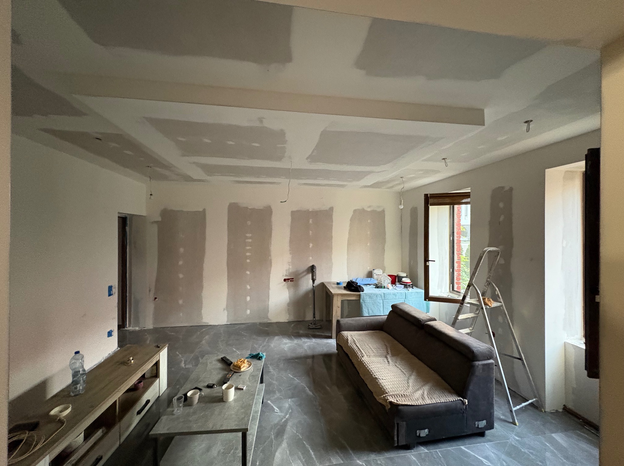 Pokój w trakcie prac wykończeniowych: ściany przygotowane do malowania, sufit z płyt karton-gips, widoczne meble i drabina. Szare płytki na podłodze.