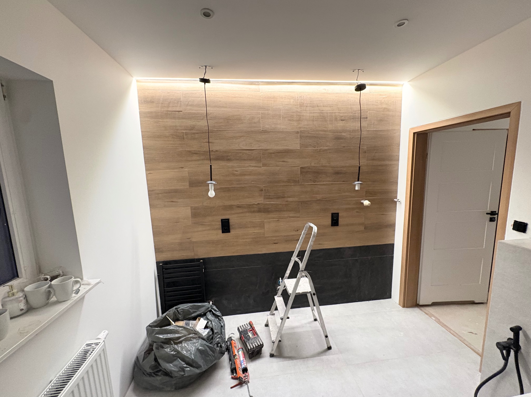 Łazienka w trakcie prac: drewnopodobne płytki na ścianie, czarne płytki poniżej, drabina, narzędzia, worki. Widoczne niedokończone oświetlenie i drzwi.