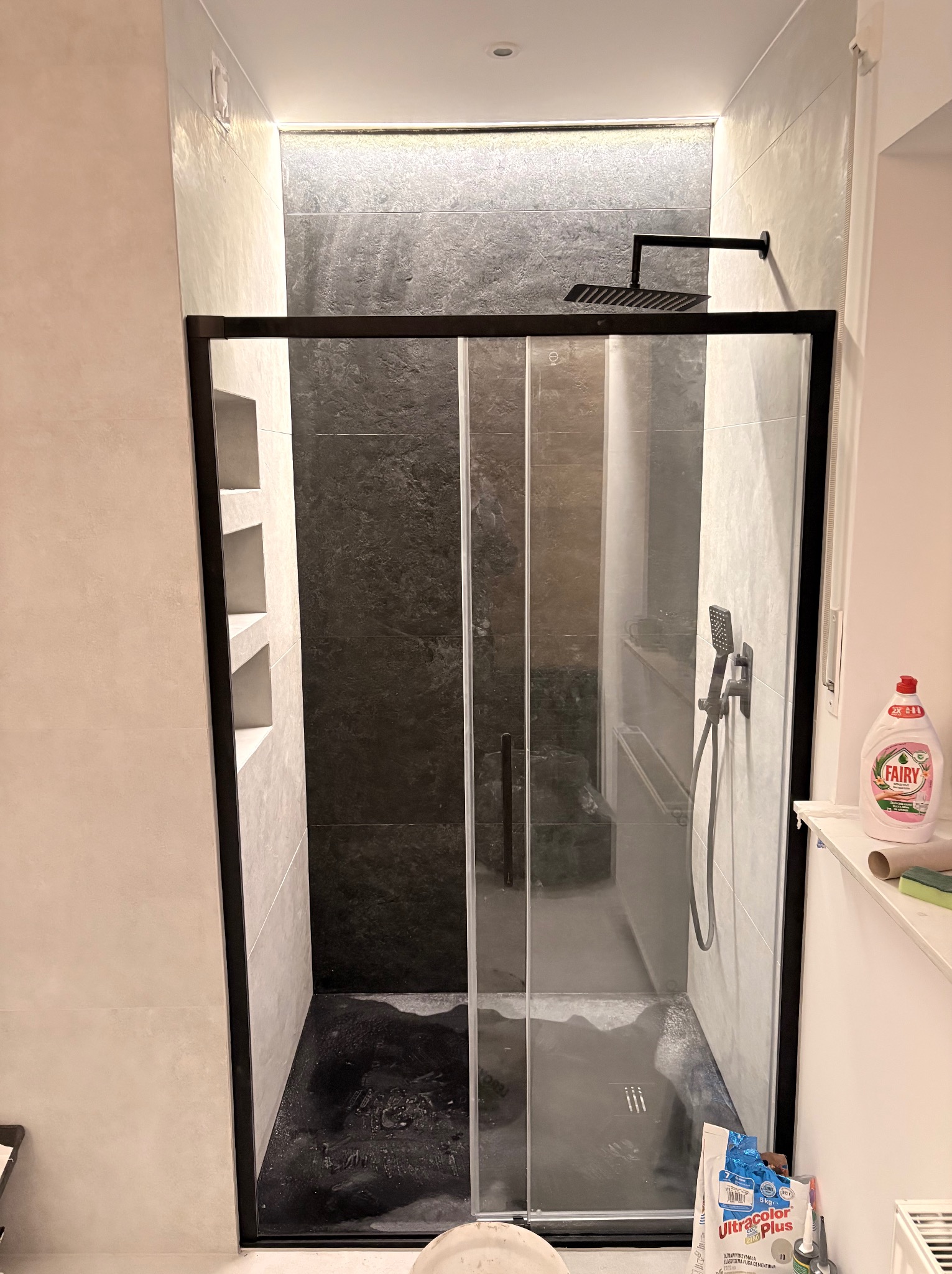 Nowoczesna kabina prysznicowa walk-in z czarną ramą i deszczownicą w wykończonej łazience. Widoczne wnęki w ścianie i butelka płynu do naczyń na półce.
