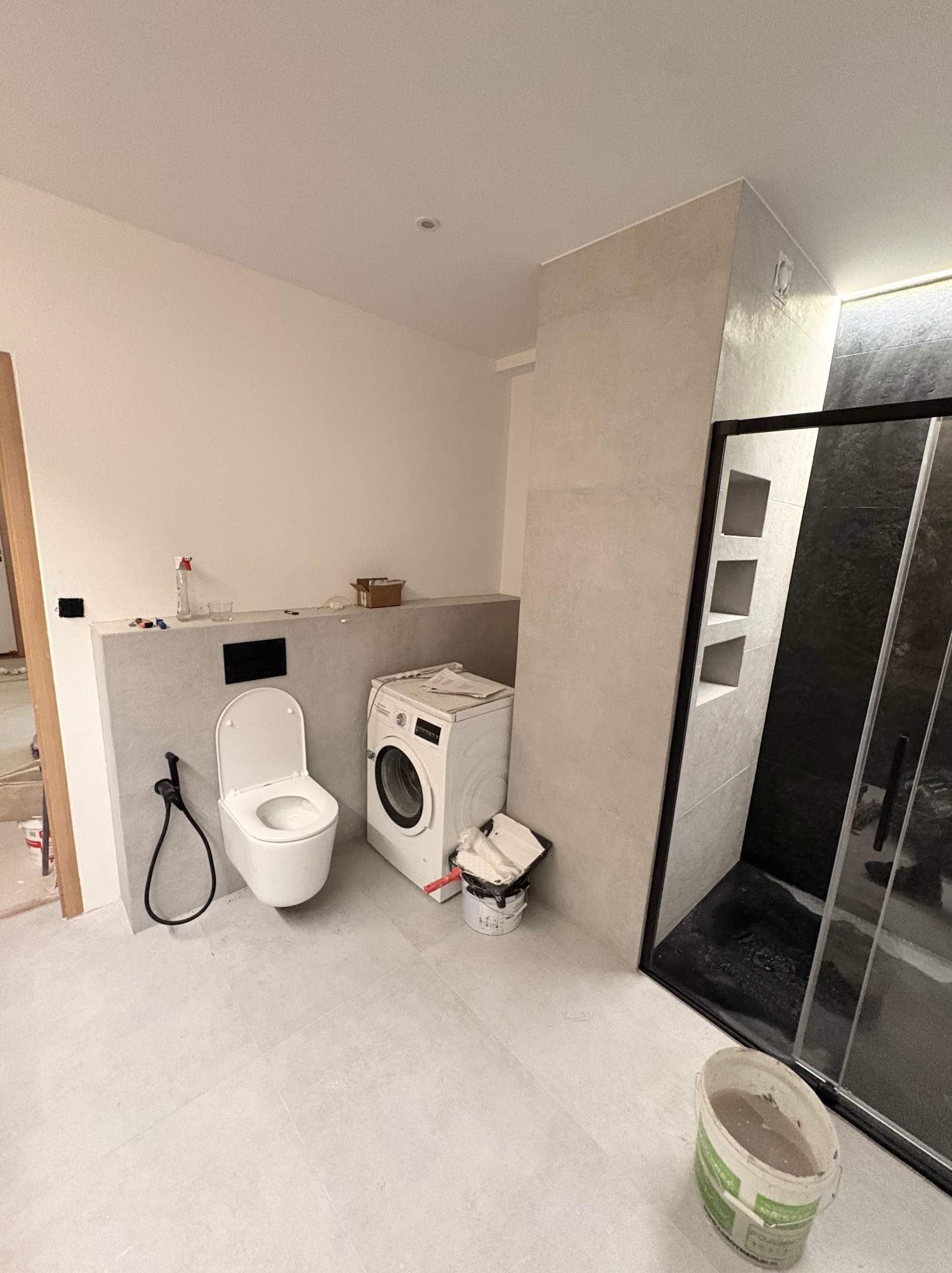 Jasna łazienka po remoncie: kabina prysznicowa z czarnymi elementami, toaleta podtynkowa, pralka. Widoczne płytki na ścianach i podłodze, oraz akcesoria łazienkowe.