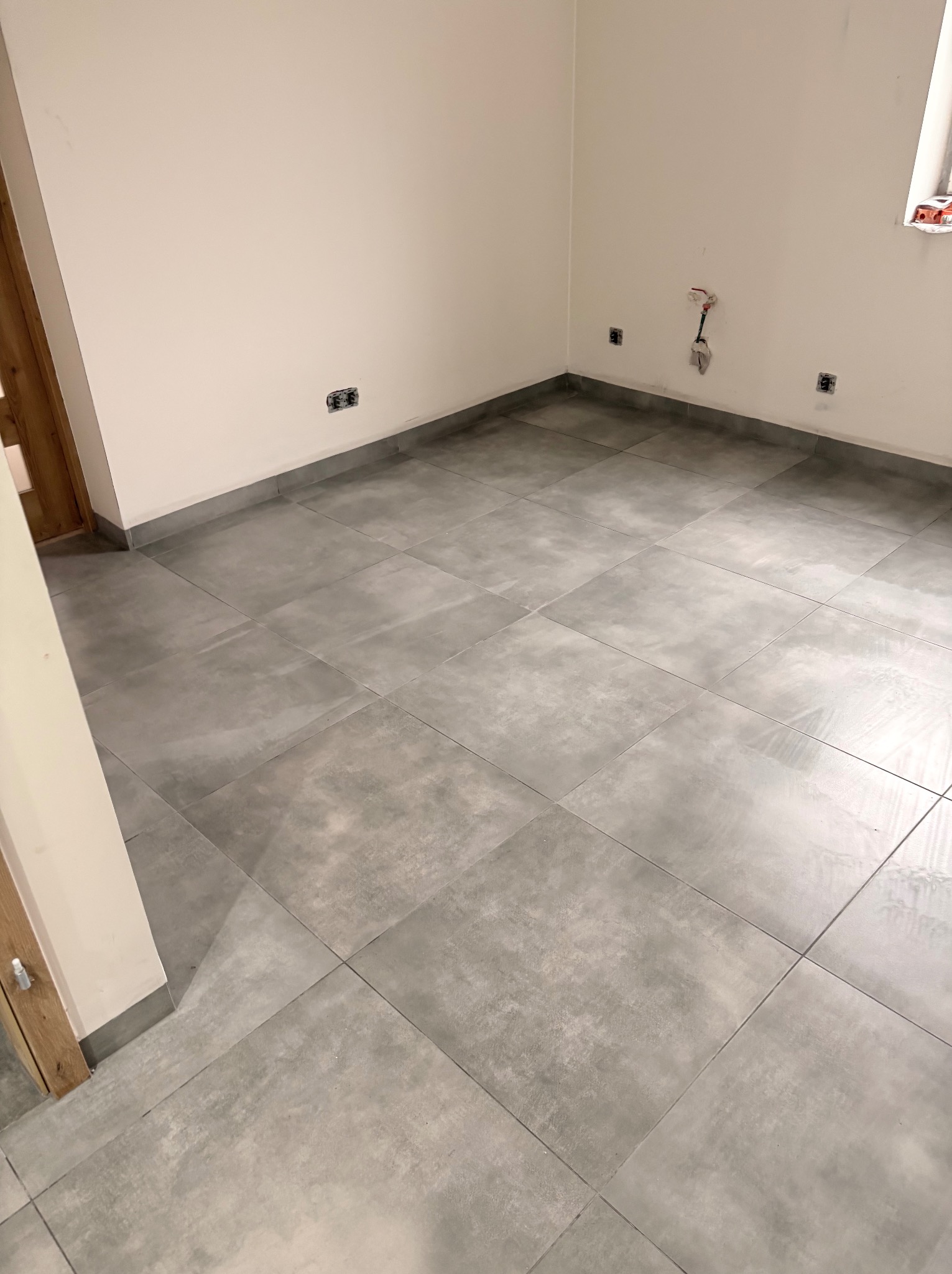 Ułożone szare płytki podłogowe w pomieszczeniu w Zabrzu. Widoczne przyłącza wodne i elektryczne na ścianie. Minimalistyczny styl wykończenia.