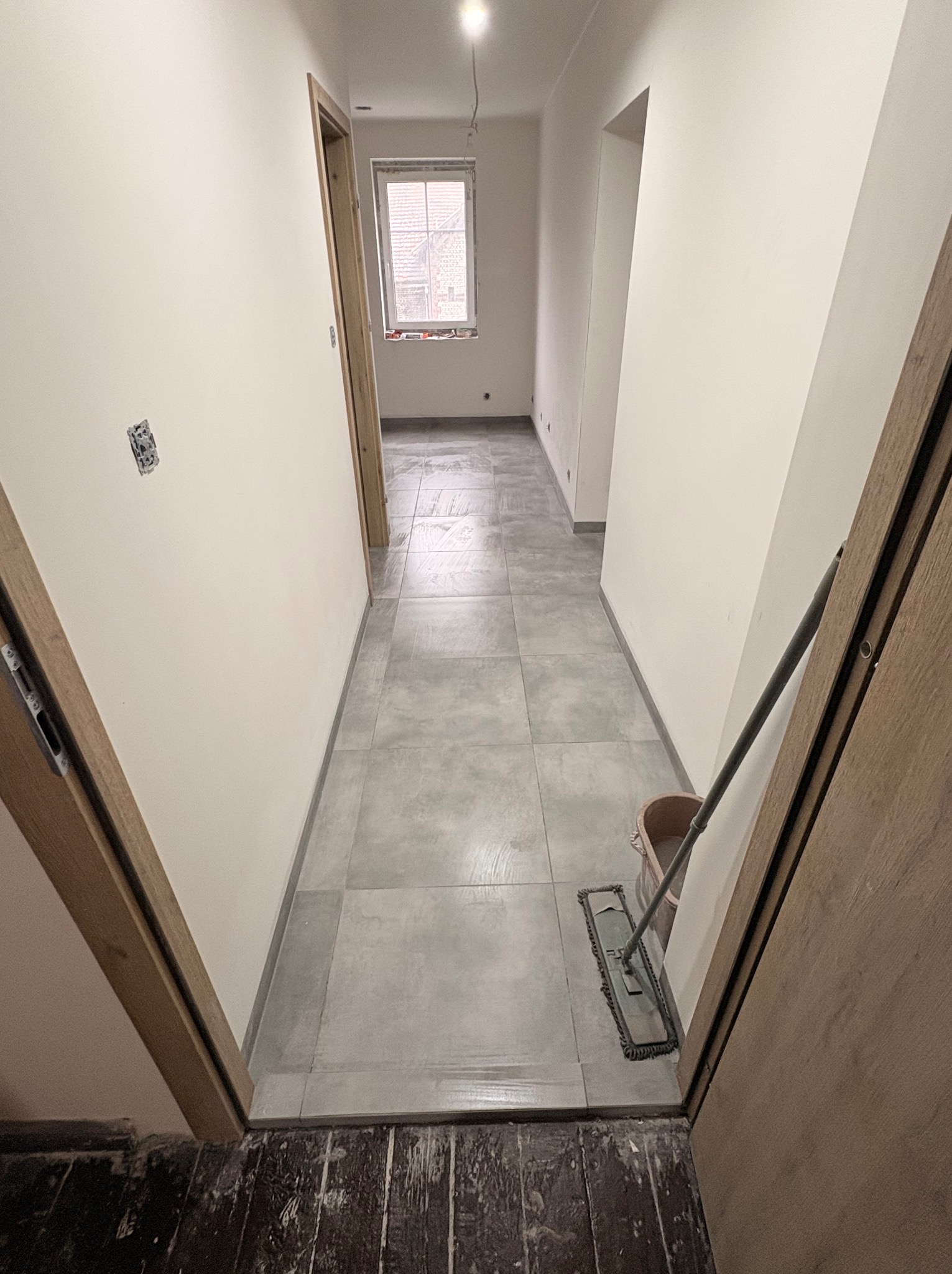 Wykończony korytarz z szarymi płytkami podłogowymi, widok z perspektywy otwartych drzwi. Ściany pomalowane na biało, mop i wiadro stoją z boku.