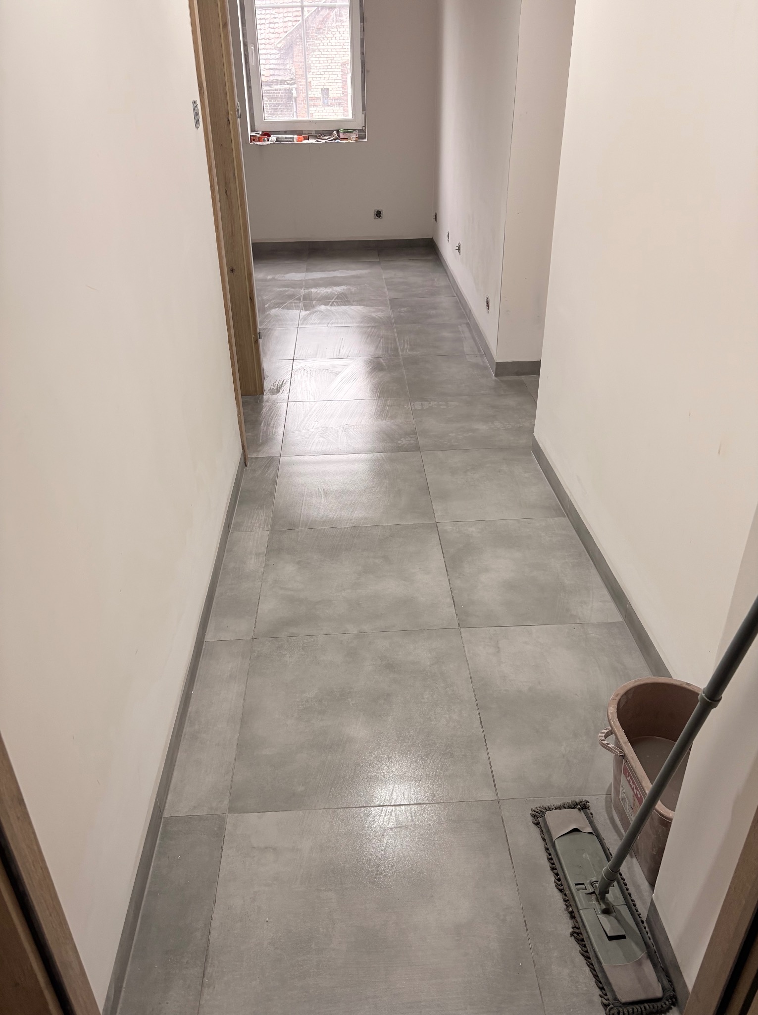 Wnętrze korytarza z szarymi, kwadratowymi płytkami podłogowymi. Ściany pomalowane na biało. Widoczny mop i wiadro. Okno w tle.
