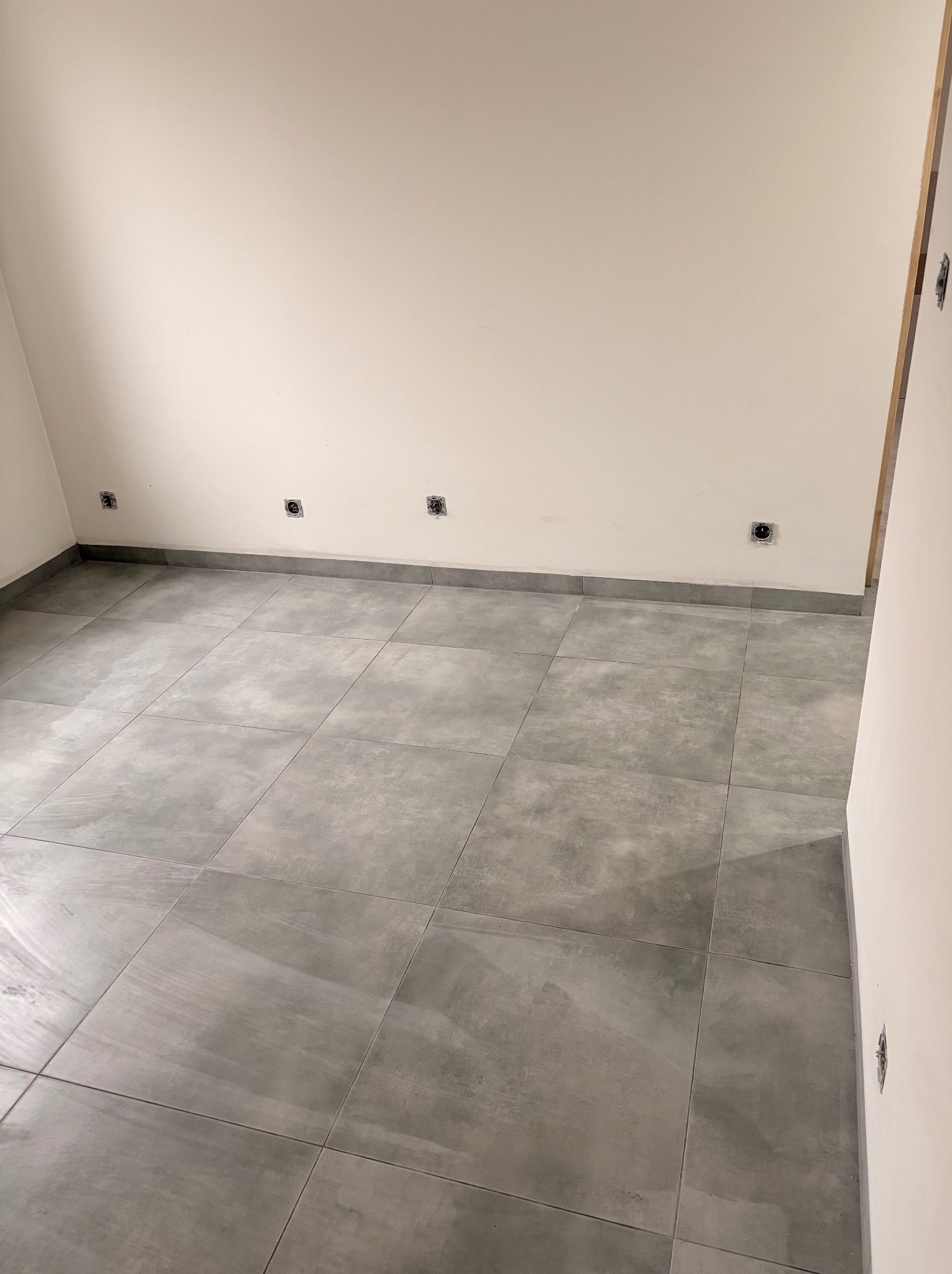 Wykończone wnętrze z szarymi, kwadratowymi płytkami podłogowymi i gładkimi, białymi ścianami z widocznymi puszkami elektrycznymi. Minimalistyczny design.