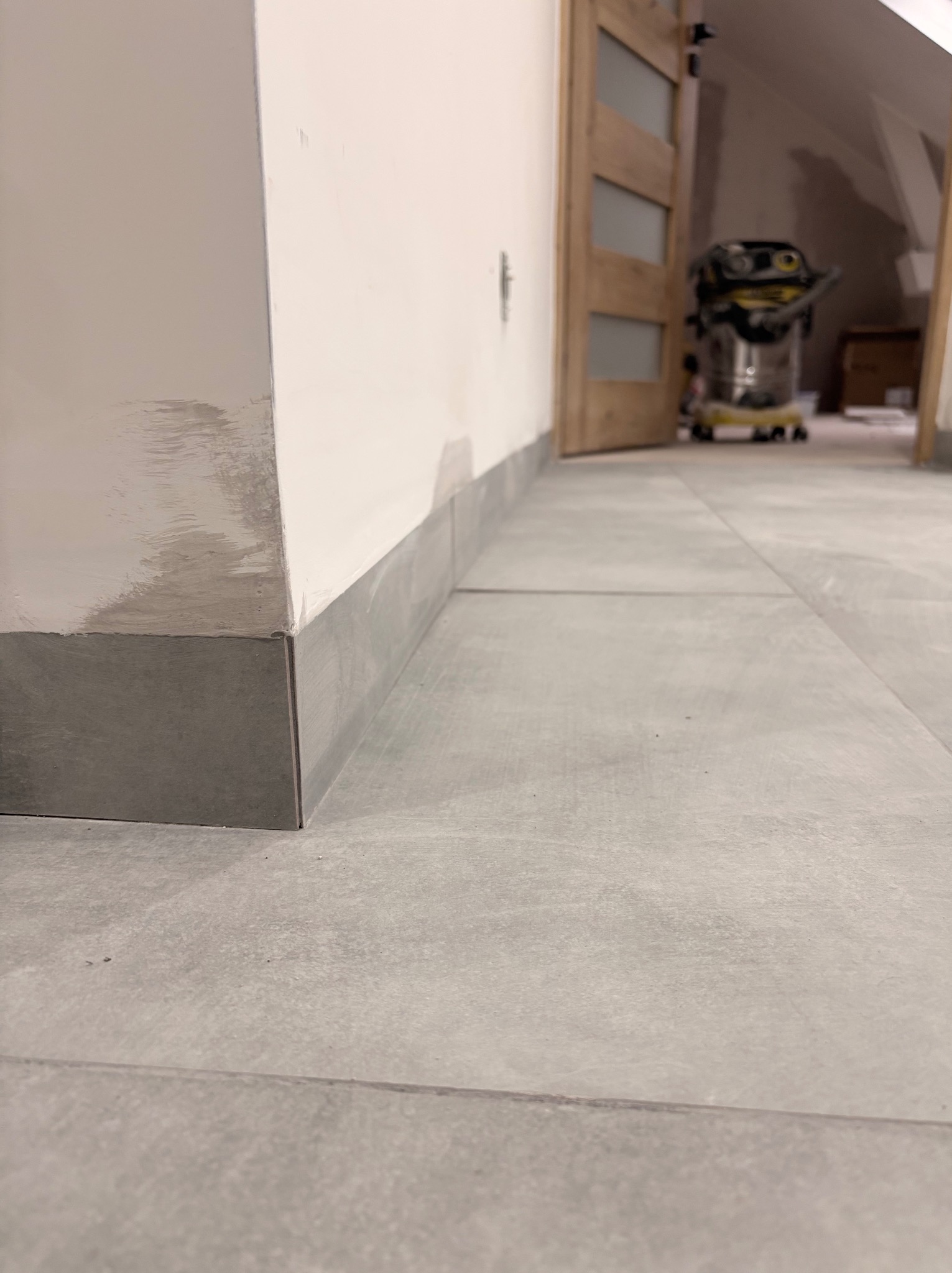 Szare płytki podłogowe i przypodłogowe w jasnym wnętrzu, widoczny fragment ściany z śladami po pracach wykończeniowych, otwarte drzwi w tle.