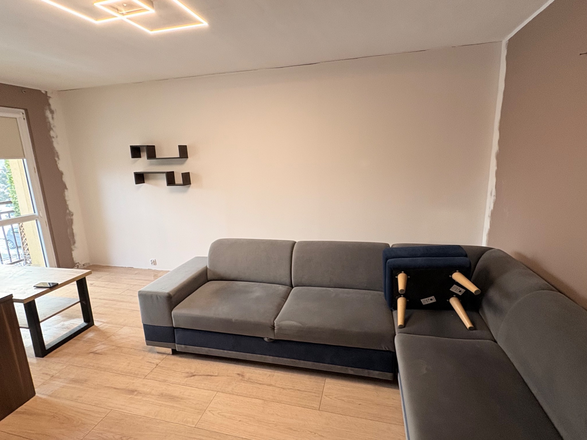 Jasne wnętrze pokoju z szarą sofą i drewnianą podłogą. Na ścianie widoczne półki. Ściany przygotowane do malowania, widoczne ślady po szpachlowaniu.