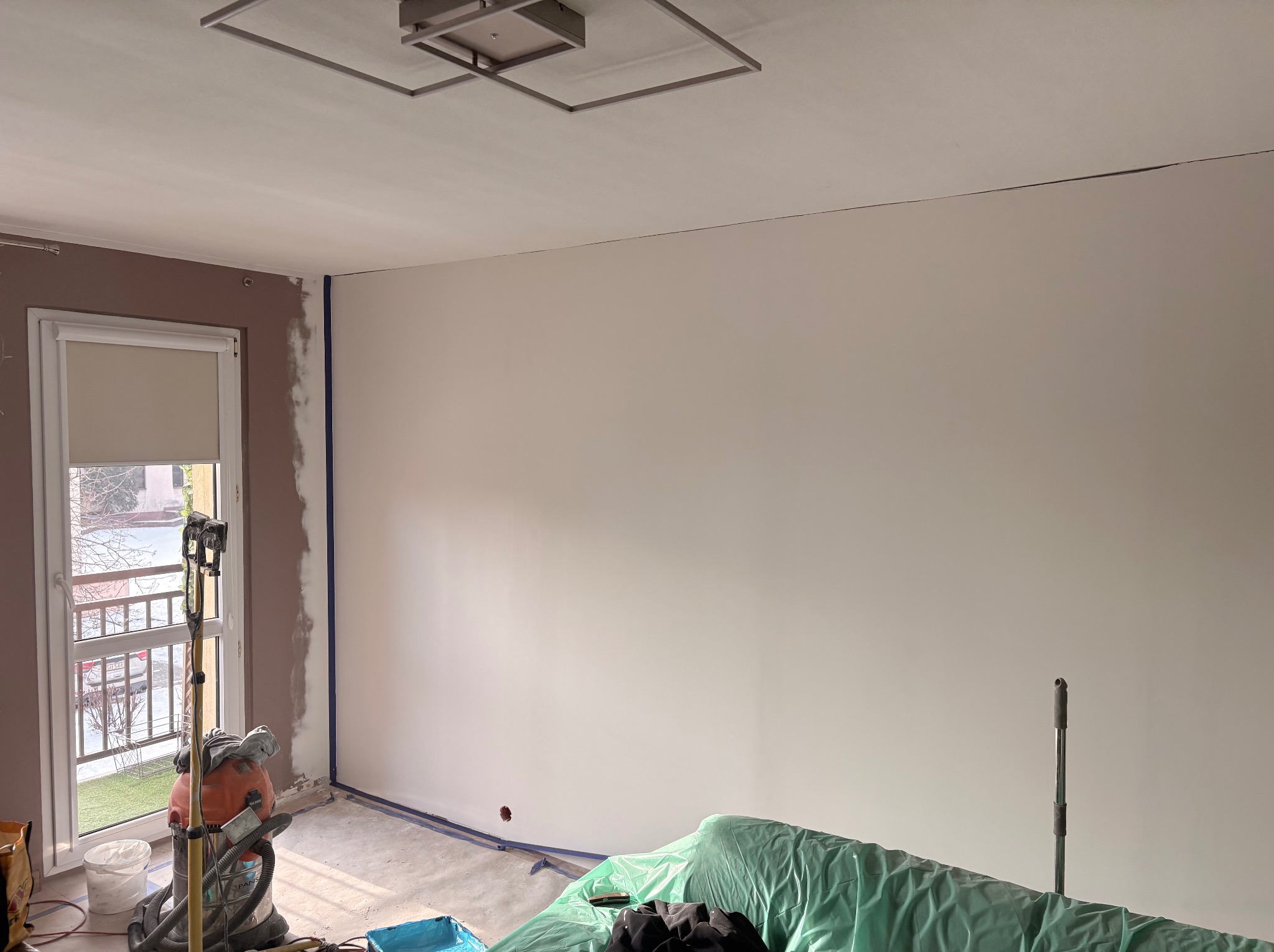 Pokój w trakcie malowania: ściana pomalowana na biało, okno z widocznymi śladami szpachlowania, zabezpieczona podłoga, odkurzacz przemysłowy. Widoczne narzędzia malarskie.