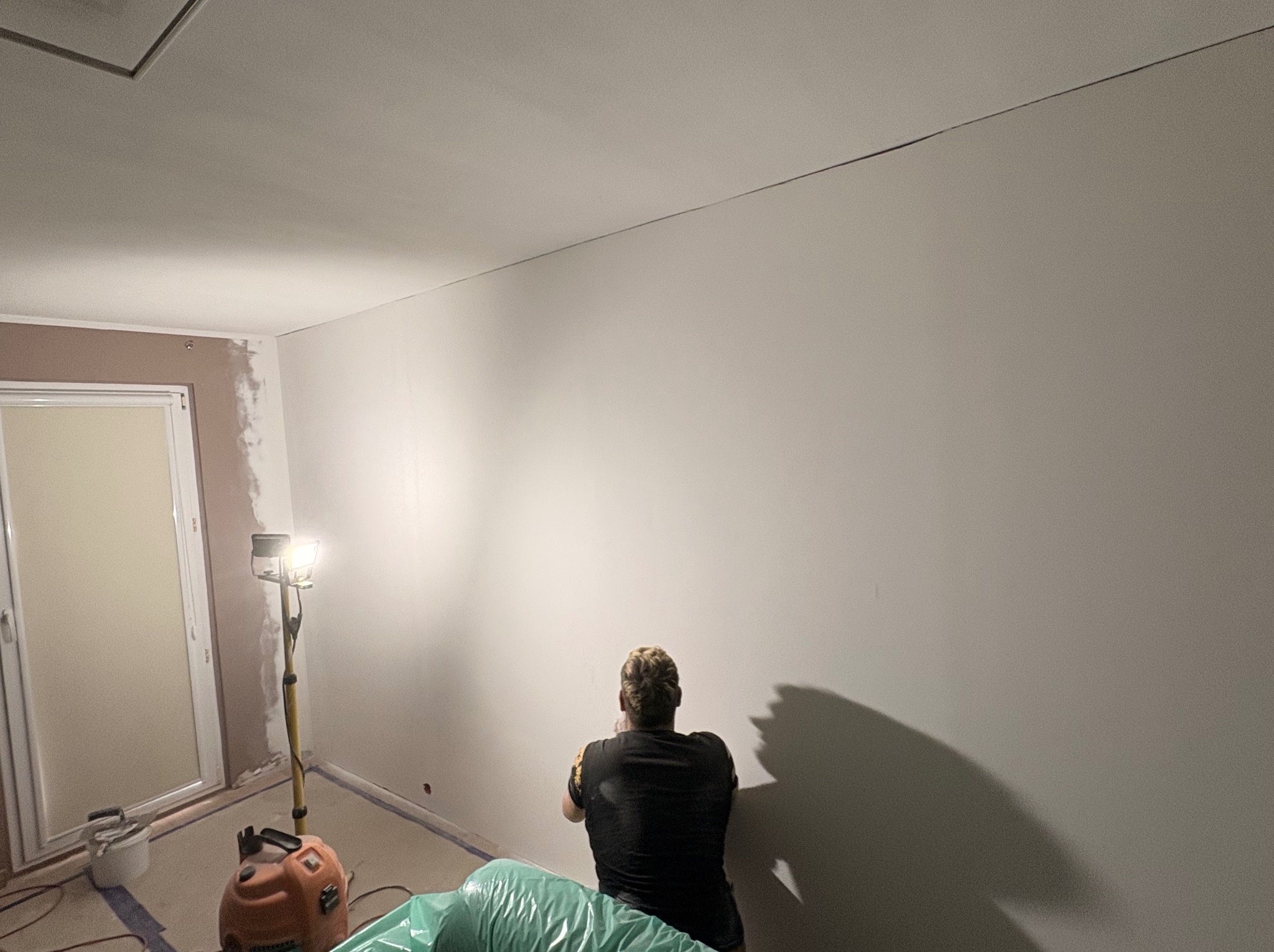 Pracownik w trakcie szpachlowania ściany w pomieszczeniu, widoczna lampa budowlana i zabezpieczenia podłogi. Ściana przygotowana do malowania.
