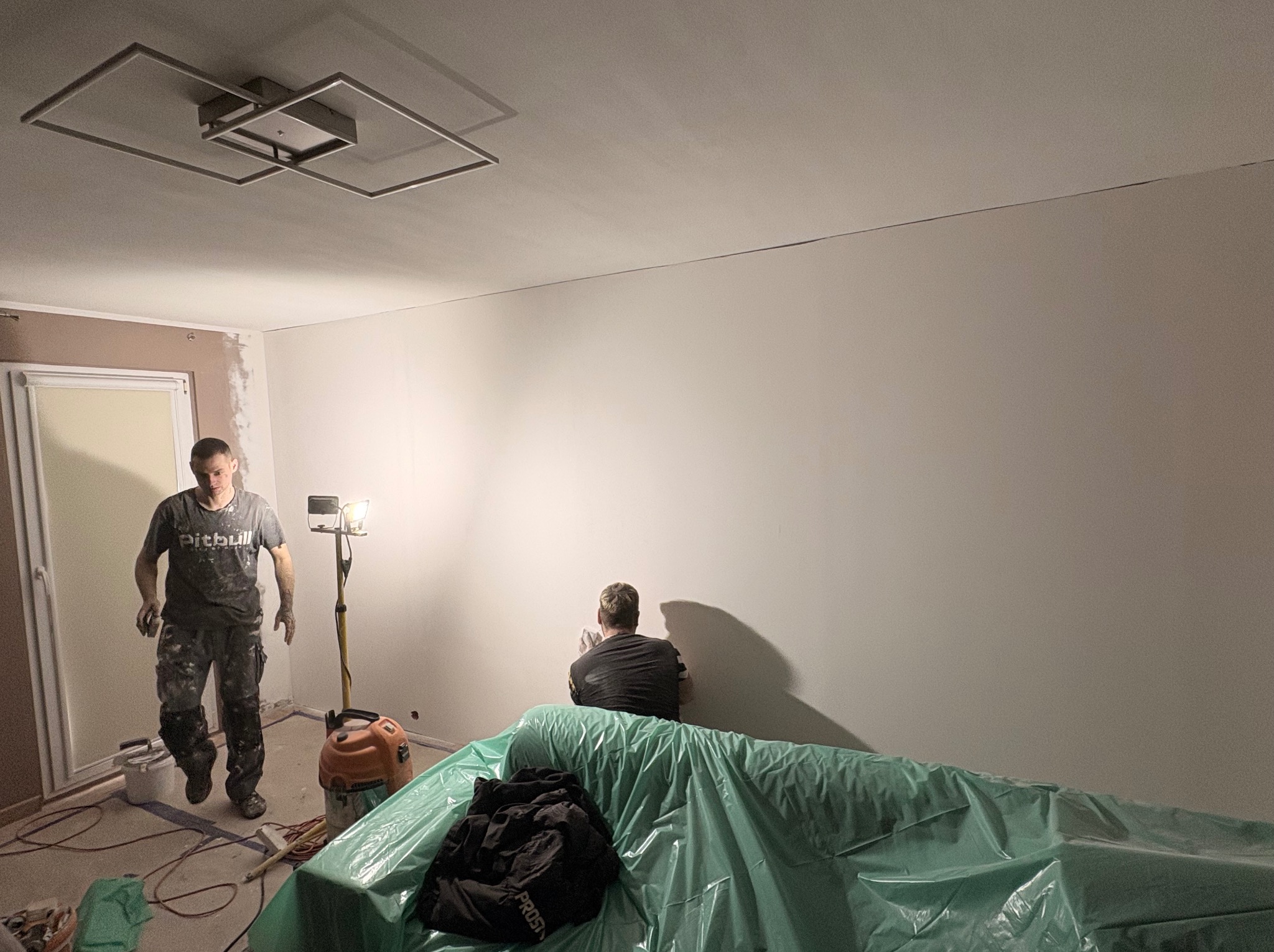 Dwóch mężczyzn podczas prac wykończeniowych w pomieszczeniu. Jeden wchodzi, drugi szpachluje ścianę. Widoczne narzędzia, lampa i zabezpieczone meble folią.