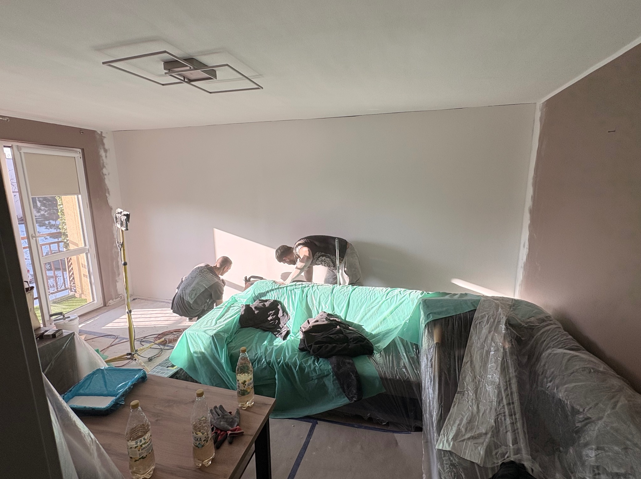 Dwóch mężczyzn szpachluje ścianę w jasnym pokoju, zabezpieczony mebel pod folią, widoczne narzędzia i oświetlenie. Ściana częściowo w kolorze.