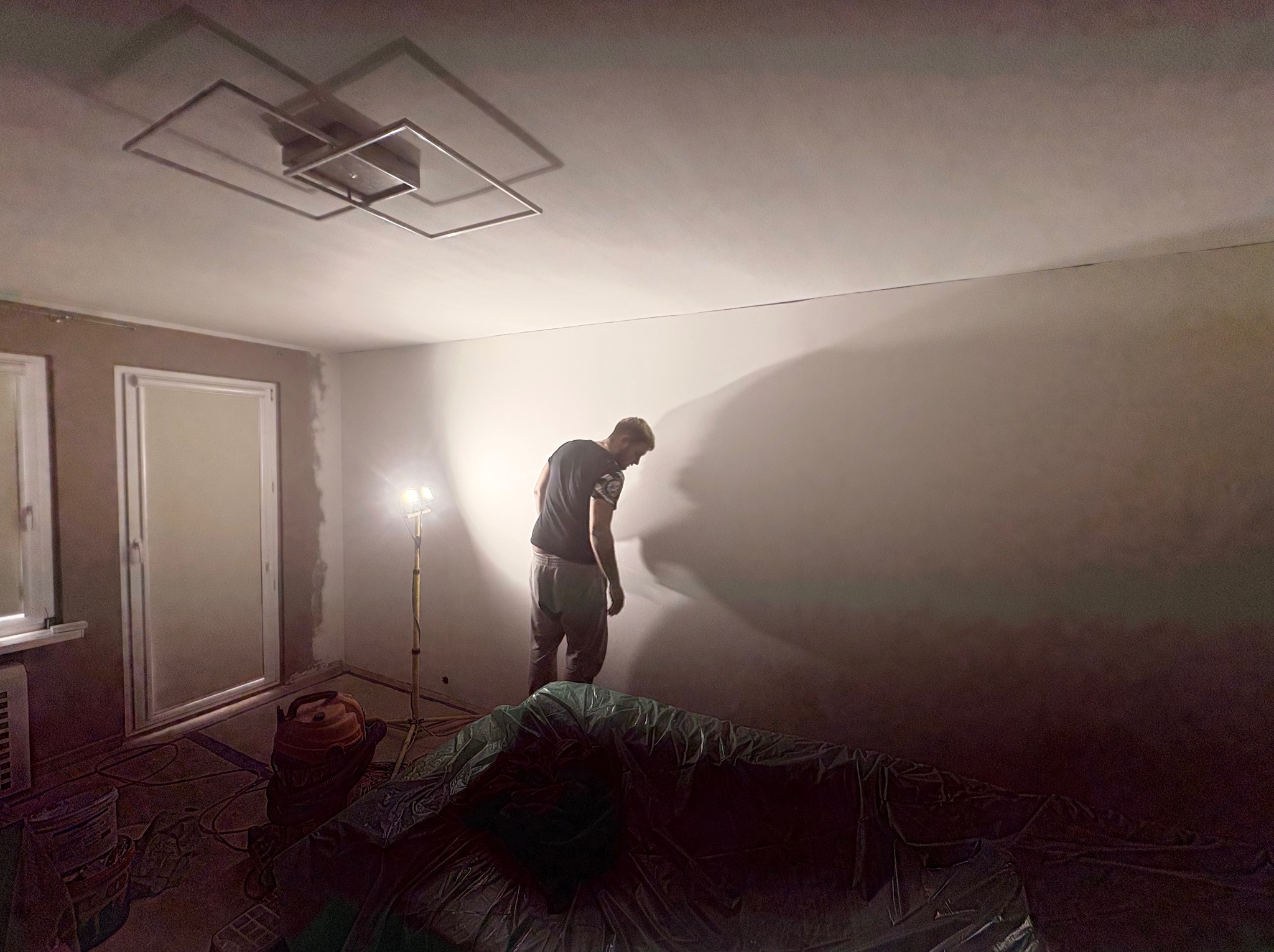Pokój w trakcie prac wykończeniowych: ściana po nałożeniu gładzi, lampa budowlana, zakryte folią meble. Widoczny pracownik w trakcie pracy.