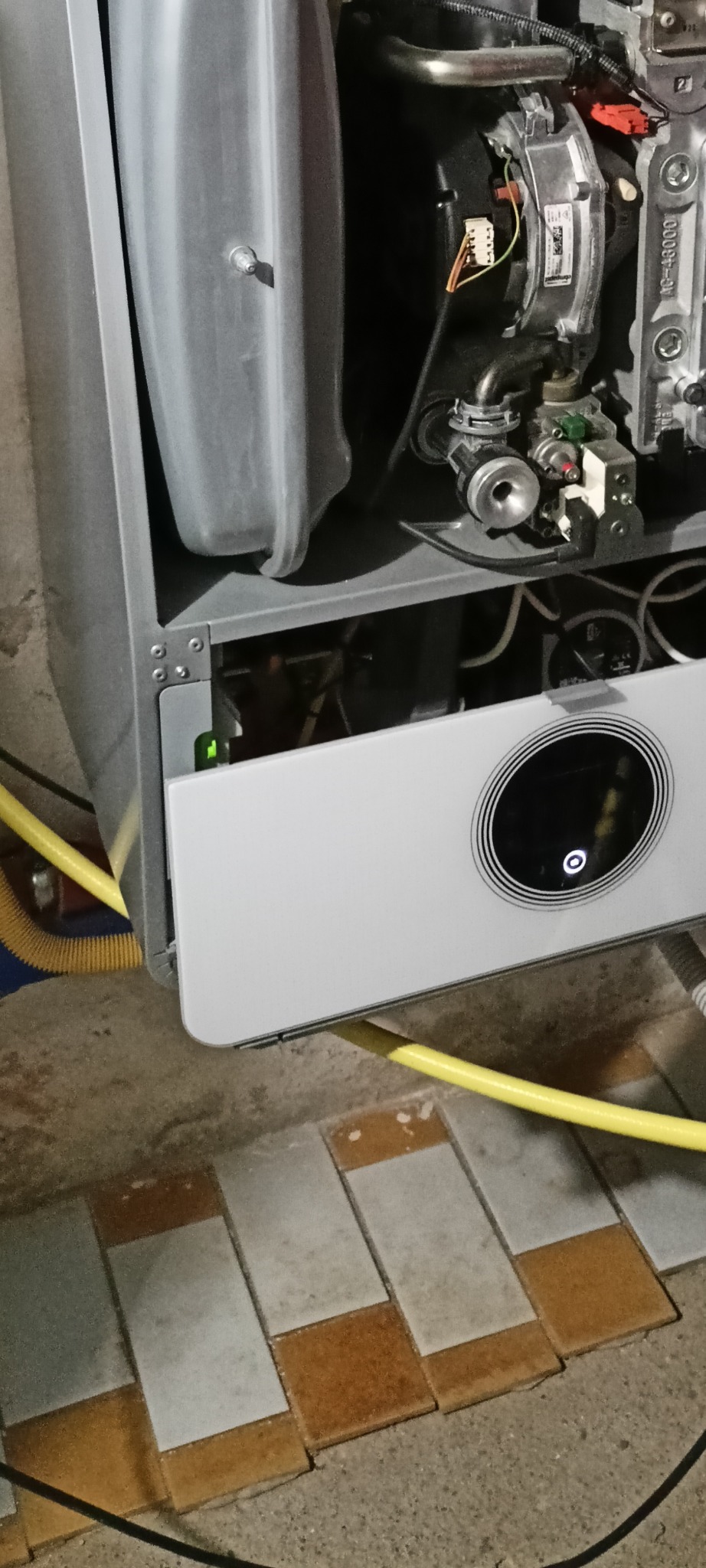 Otwarty panel kotła gazowego z widocznymi elementami wewnętrznymi i żółtym przewodem, na tle płytek podłogowych. Widok z bliska na wnętrze urządzenia.