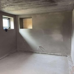 Łukasz Krawczyk - Surowe wnętrze po tynkowaniu: szare ściany z widocznymi przewodami, okno z tkaniną i surowy betonowy sufit. Minimalistyczny, industrialny klimat.