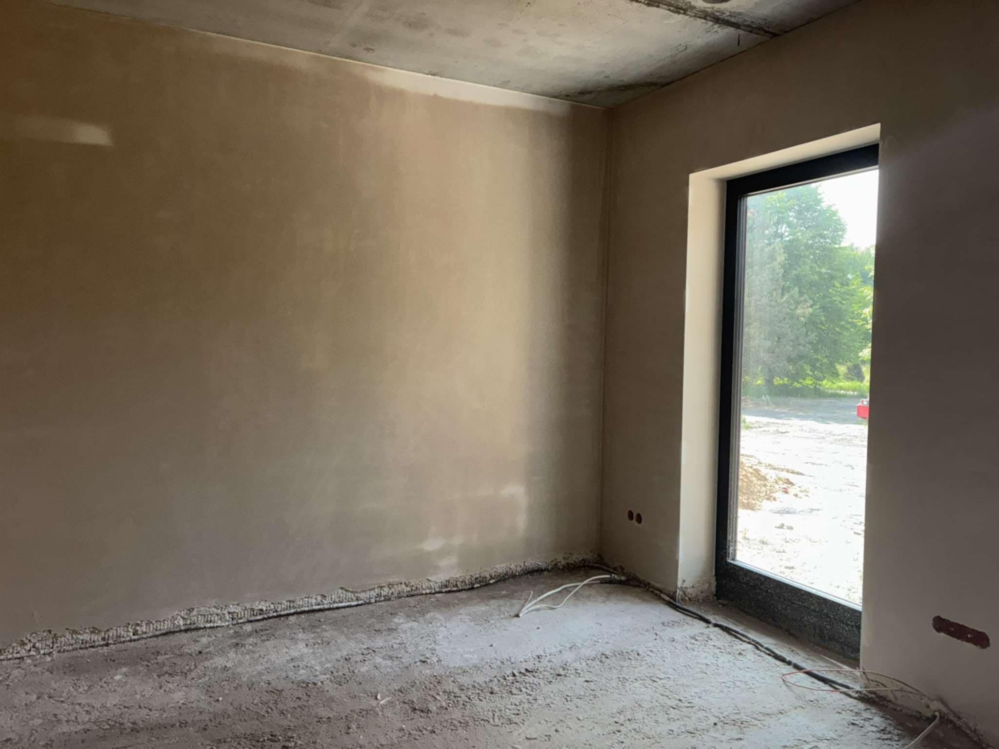 Pokój po tynkowaniu maszynowym: gładkie, beżowe ściany, surowy betonowy sufit, okno z widokiem na zieleń. Widoczne przewody elektryczne przy podłodze.