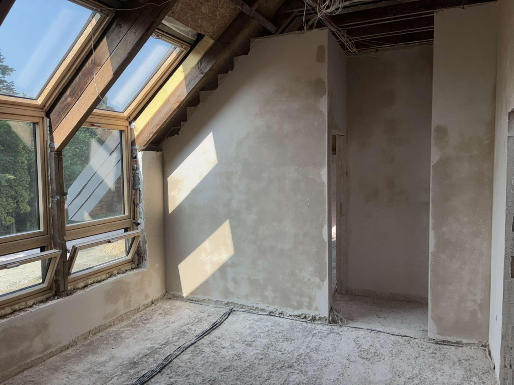 Poddasze po szpachlowaniu ścian, widoczne okna dachowe i surowe wykończenie podłogi. Ściany przygotowane do dalszych prac wykończeniowych, z widocznymi śladami szpachli.