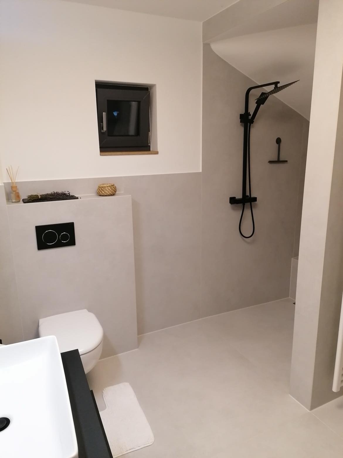 Minimalistyczna łazienka w Wilczkowicach z czarną armaturą, małym oknem i beżowymi ścianami. Umywalka z czarnym blatem, toaleta podtynkowa i kabina typu walk-in.