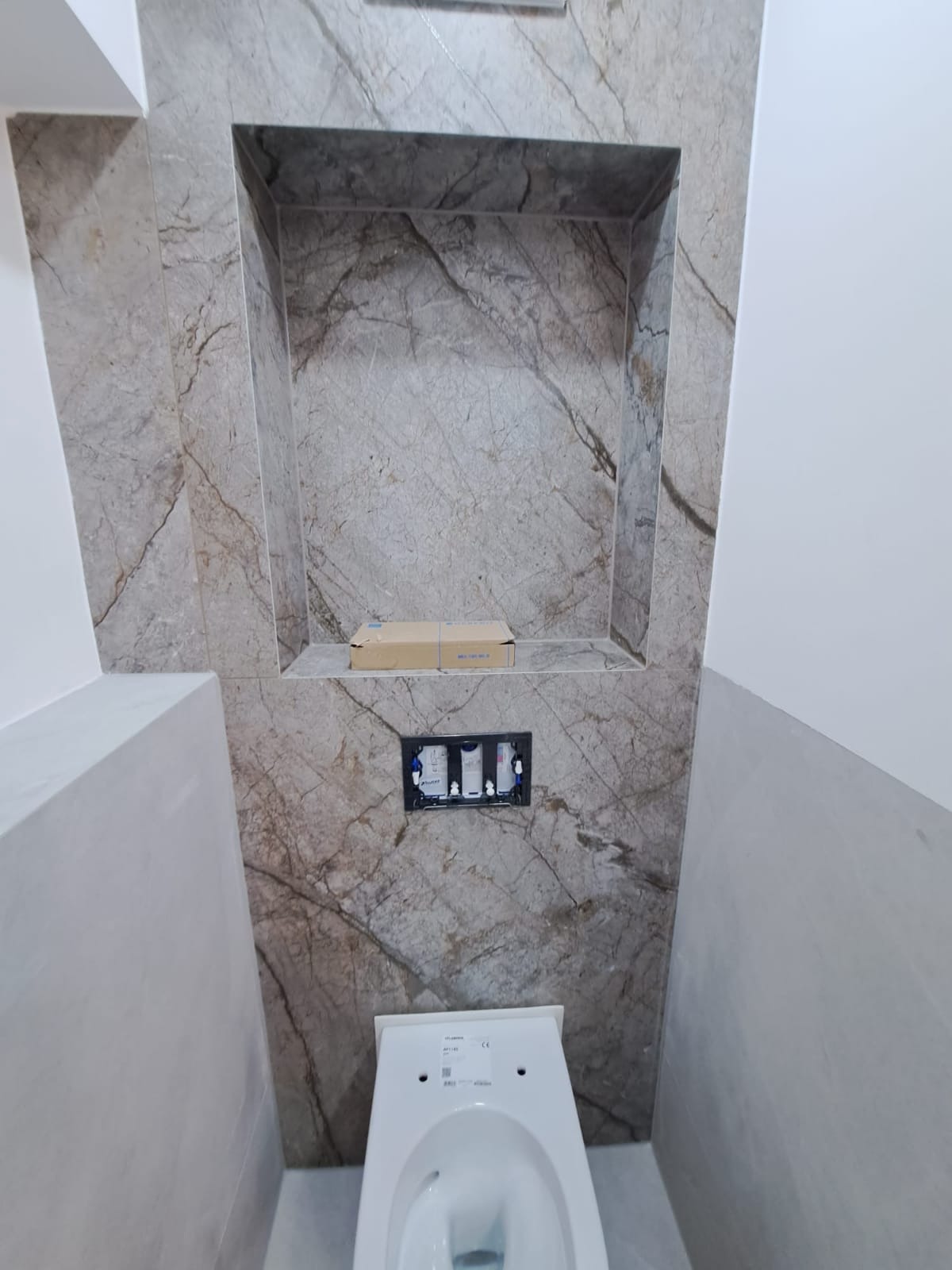 Wykończona toaleta z płytkami imitującymi marmur, wnęka w ścianie i podtynkowy system spłukiwania. Widok z dołu, podkreślający detale wykończenia.