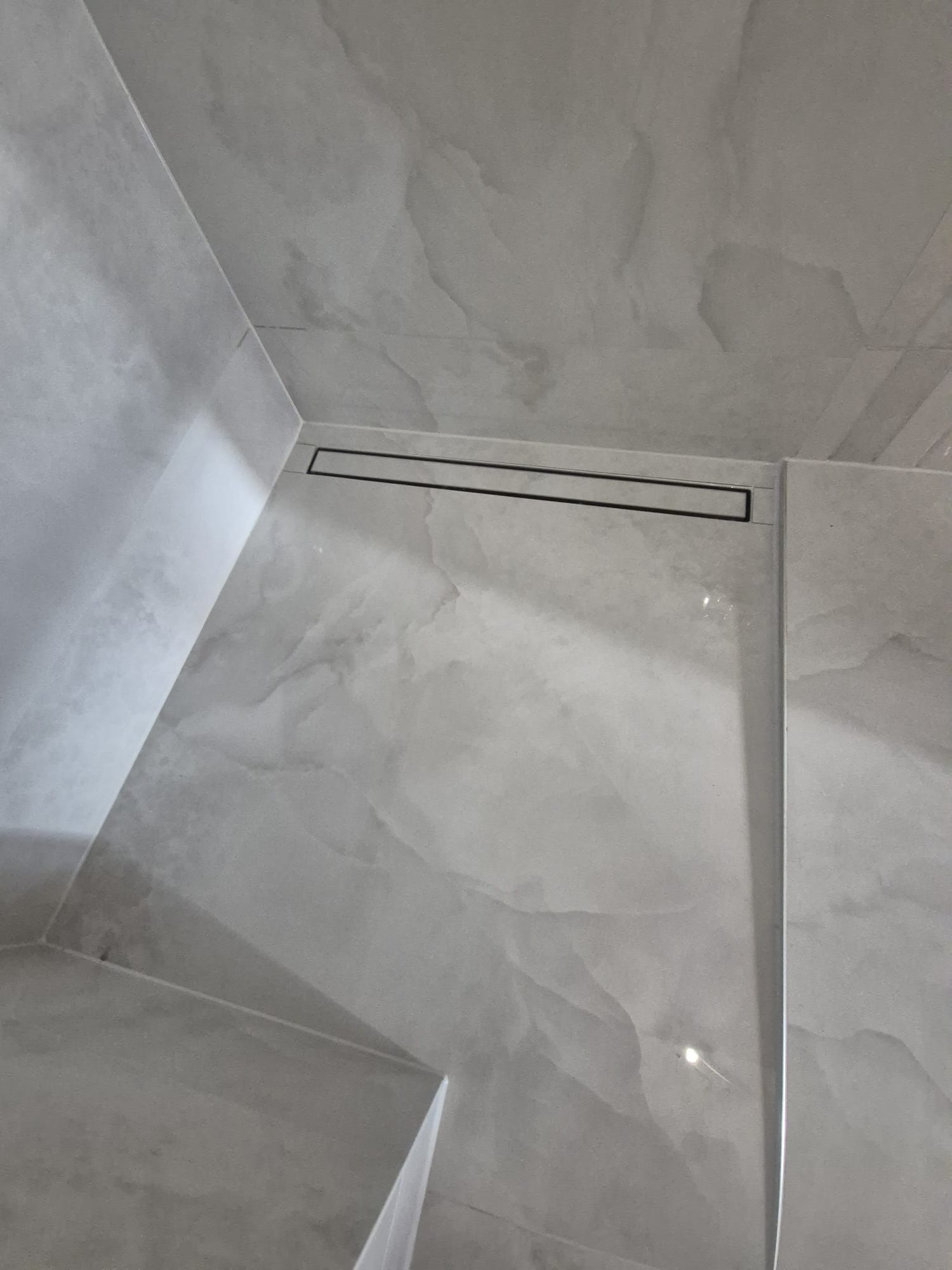 Wykończona łazienka z szarymi płytkami imitującymi marmur i czarnym odpływem liniowym. Minimalistyczny design, nowoczesne wykończenie.