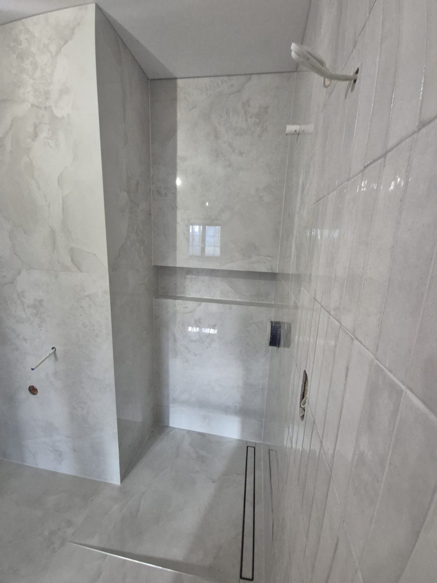 Wykończona łazienka z szarymi płytkami imitującymi marmur, widoczny odpływ liniowy i wnęka w ścianie. Minimalistyczny design, surowy styl.